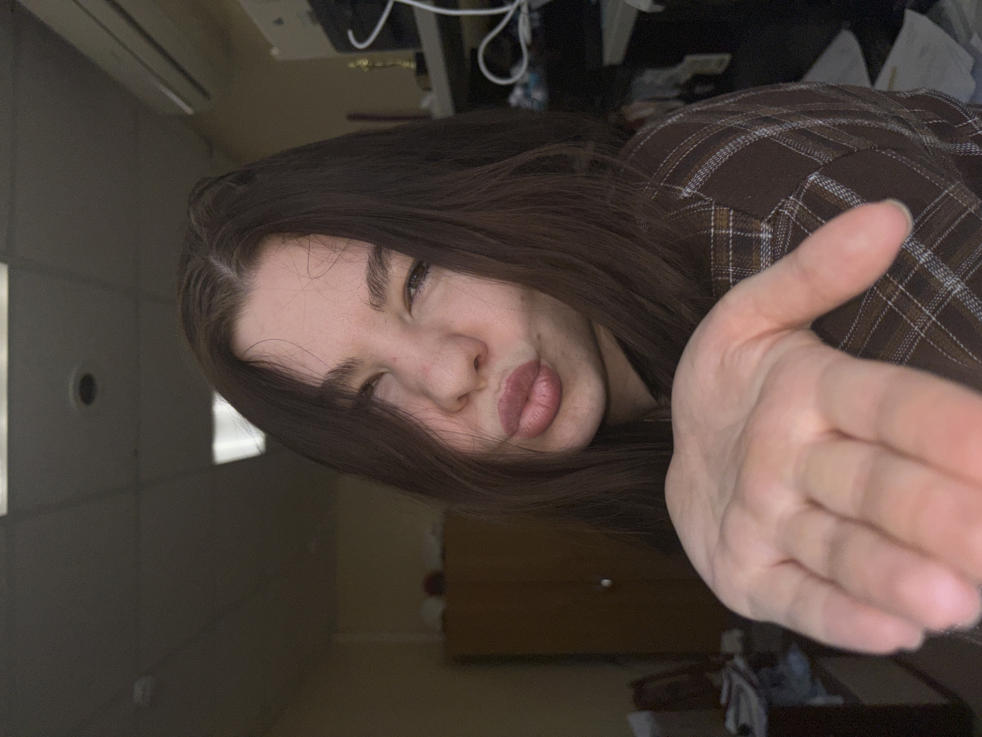 A woman with long brown hair is the main subject, posed to the side, appearing to blow a kiss or wave at the viewer. She has a brown plaid shirt. The location seems to be indoors, in what looks like an office or living space. A light-colored wall with a circular object (possibly a sensor) and a light fixture are visible in the background. Further in the background, are some office equipment and furniture are discernable, suggesting a cluttered area. It is not possible to identify the location as Perm, Russia from the visual information alone.