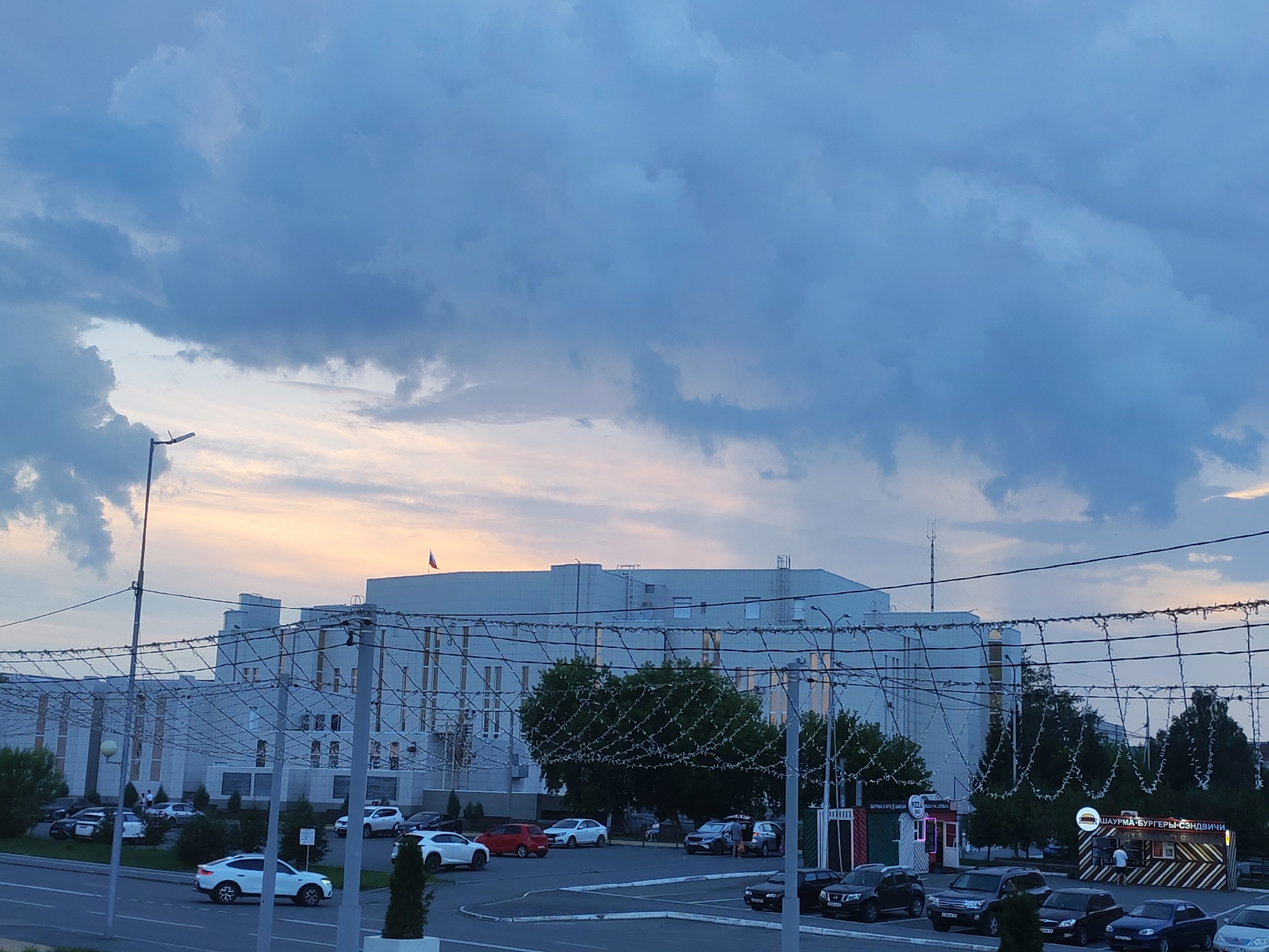 The image depicts a street scene in front of a large, modern building with a Russian flag flying on its roof. The building is light-colored and features numerous windows. In the foreground, there are several parked cars on a street and in a parking lot. Some of the cars appear to be white SUVs. A few trees are visible, and some of them are decorated with string lights. Two small food kiosks are also present in the parking lot. One kiosk has the text "PIZZA" visible, and the other has text that appears to be in Russian, possibly advertising shawarma, burgers, and sandwiches.
The sky is cloudy, with a mixture of dark blue and lighter, warmer tones suggesting either sunrise or sunset. A street lamp is visible on the left side of the image. The overall scene appears to be a relatively calm urban environment. There are a few people visible near the kiosks and one person standing near a parked car. The time of day is likely early morning or late evening due to the sky's coloration and the presence of artificial lighting on the trees. No specific location is indicated, but the Russian flag suggests a location within Russia.