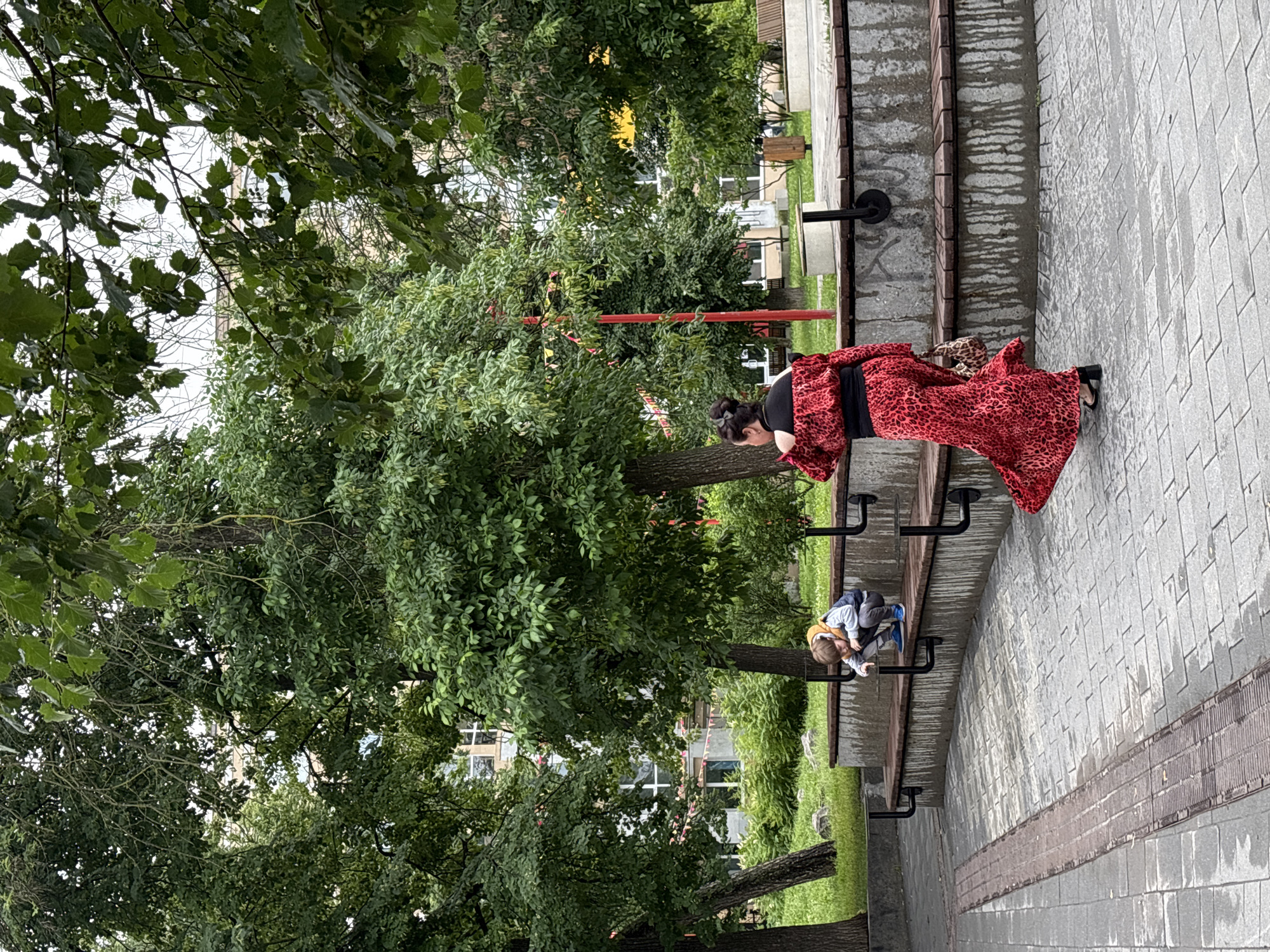In Nizhny Novgorod, Russia, on a paved area adjacent to a park with trees and buildings, a child wearing a yellow hooded jacket, grey vest, and blue sneakers is crouching on a wooden bench. They are looking down at something on the ground. Nearby, a woman in a red leopard-print dress with black accents walks away from the camera, carrying a patterned handbag. The environment includes a concrete structure with wooden seating, a sidewalk with a metal grate, and greenery.