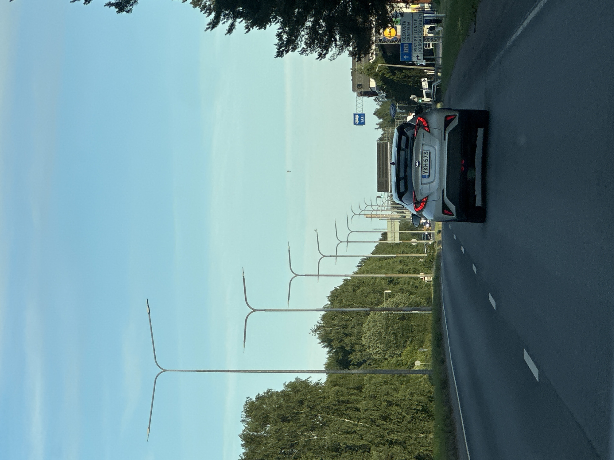 The image shows a view from a car driving on a road. The perspective is from the passenger seat looking forward and slightly to the right. The road is asphalt with white lane markings. On the left side of the road, there is a row of tall, modern lampposts with a distinctive "Y" shape at the top. Beyond the lampposts, there is a dense line of green trees. The sky is a clear, pale blue, suggesting a daytime scene, likely morning or afternoon, with good weather.
In the distance, on the right side of the road, a white car is visible. It is a modern hatchback with its rear facing the camera. The license plate of this car is visible and reads "YKH 573". There are also traffic signs visible in the distance, indicating road directions and possibly services. One sign has text that appears to include "120 KEHRUSTA CENTRUM" and "RISTIPELLONTIE KORSAKERSVÄGEN". Another sign in blue has "TAXI" written on it.
The scene is an outdoor, possibly semi-urban or suburban environment, given the presence of lampposts, road signs, and the general appearance of the surroundings. There are no people clearly visible. The overall mood is neutral, a snapshot of a typical journey. The image is oriented vertically, requiring rotation for a standard view.