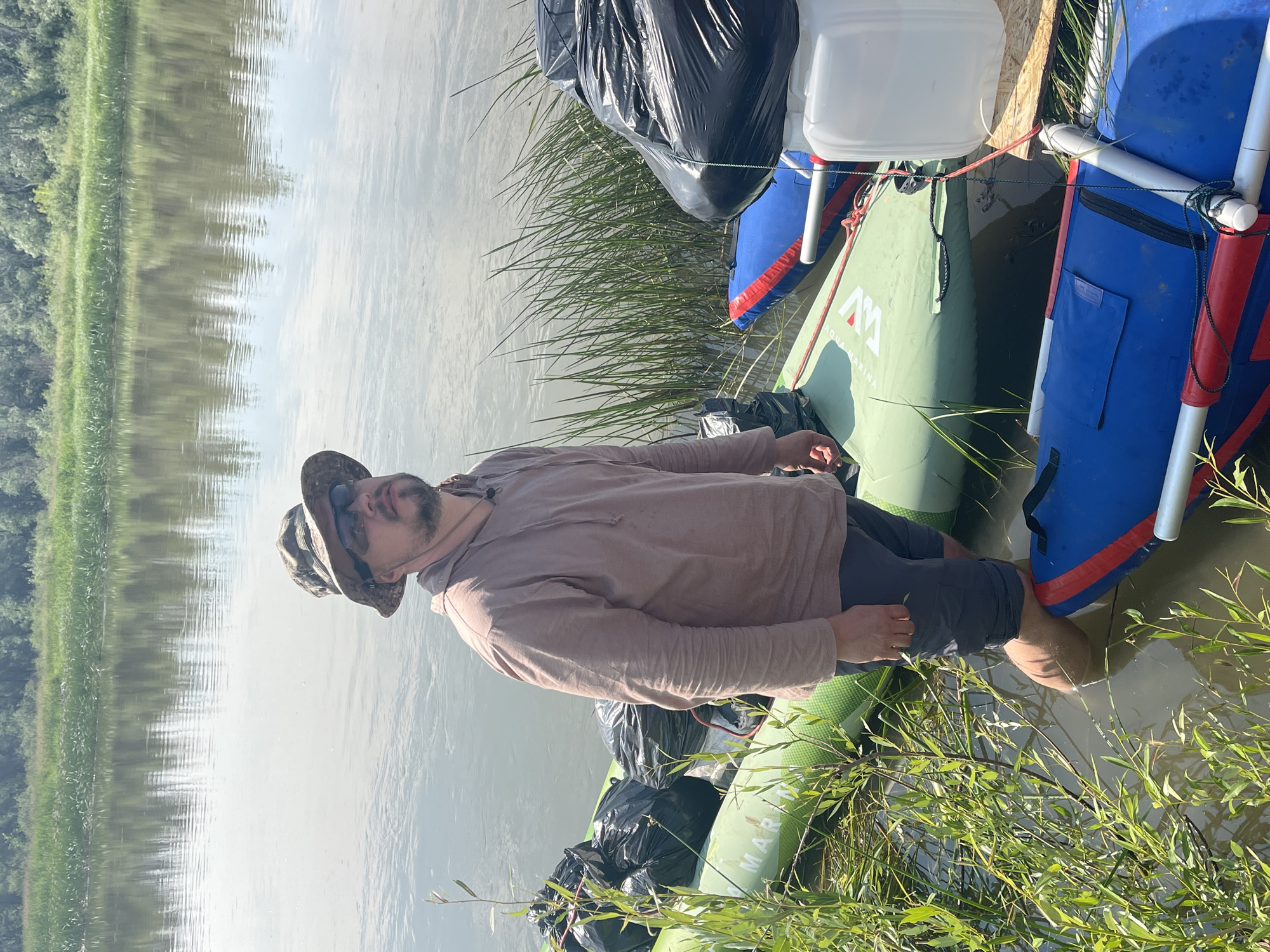 A man stands in shallow, muddy water next to several inflatable rafts loaded with black trash bags. He is wearing a camouflage hat, sunglasses, a light brown hoodie, and dark shorts. The rafts are green and blue. In the background, a calm river reflects the sky and a dense line of green trees. The scene appears to be a riverbank in a rural or natural setting, potentially near Nezhinka, Russia, given the context.