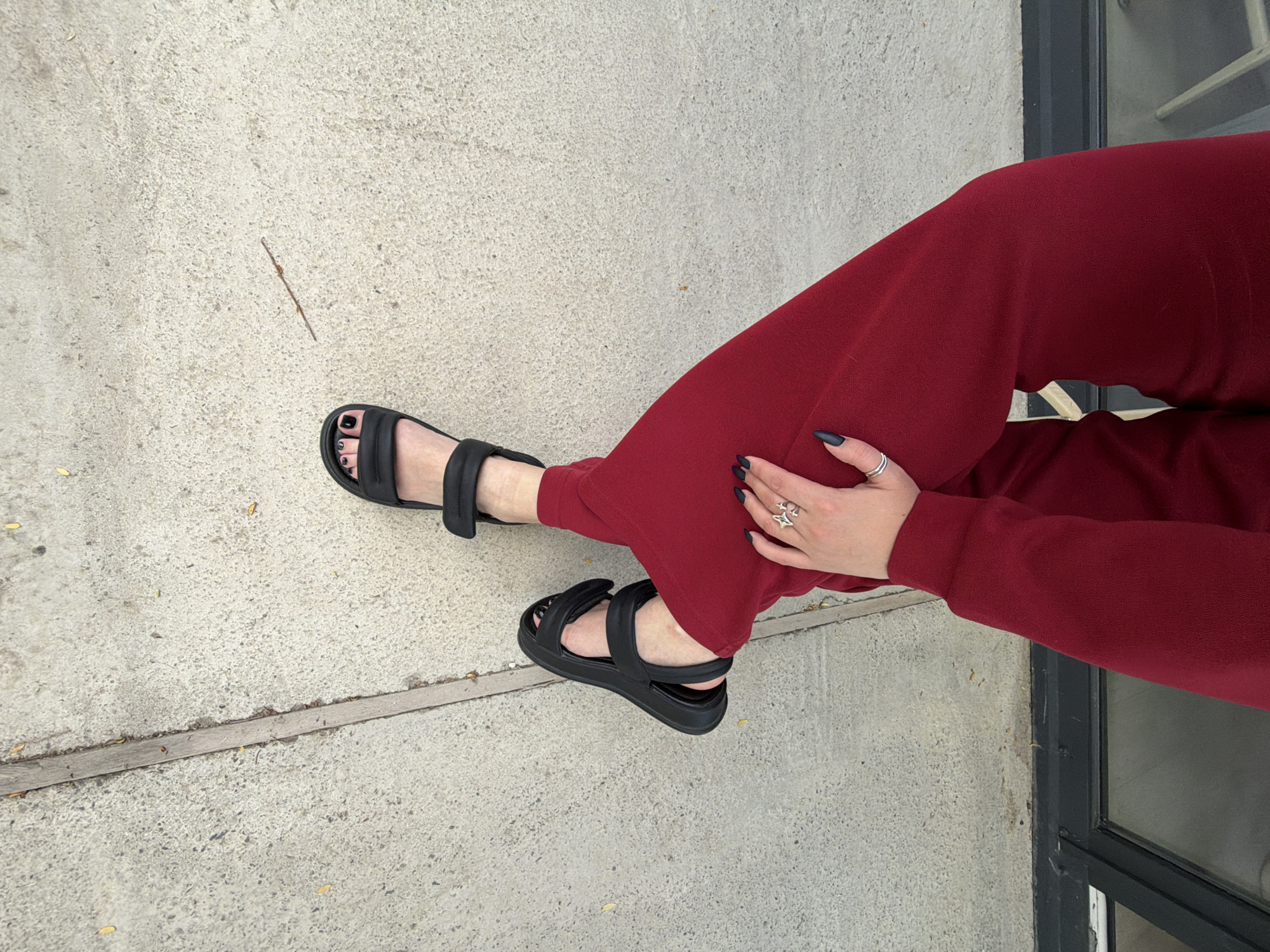 A photo shows a person's lower body wearing red pants and black sandals, set against a concrete background and a portion of a doorway. The person's legs are crossed, with the right leg positioned over the left. On their left hand, visible rings are worn. The concrete surface shows a long, thin object, and there's a portion of a door or window frame visible in the background. The overall scene appears casual and unplanned, possibly captured outdoors in an urban setting.