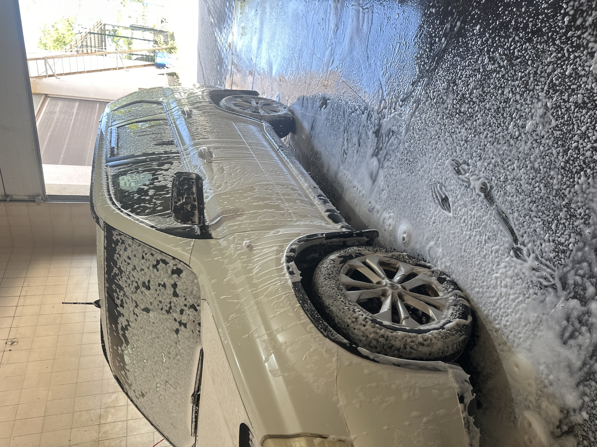 In Shatava, Ukraine, a white SUV is being washed at an outdoor car wash. The vehicle is covered in soap suds, which are also present on the ground. The car wash appears to be located under an overhang, with an open garage door in the background. Sunlight streams in from the open door, illuminating the scene. The ground is wet and reflective, with tire tracks visible in the suds. A ladder can be seen outside the garage, suggesting a multi-story structure or work area. The overall impression is of a routine car cleaning taking place in a well-lit, functional environment.