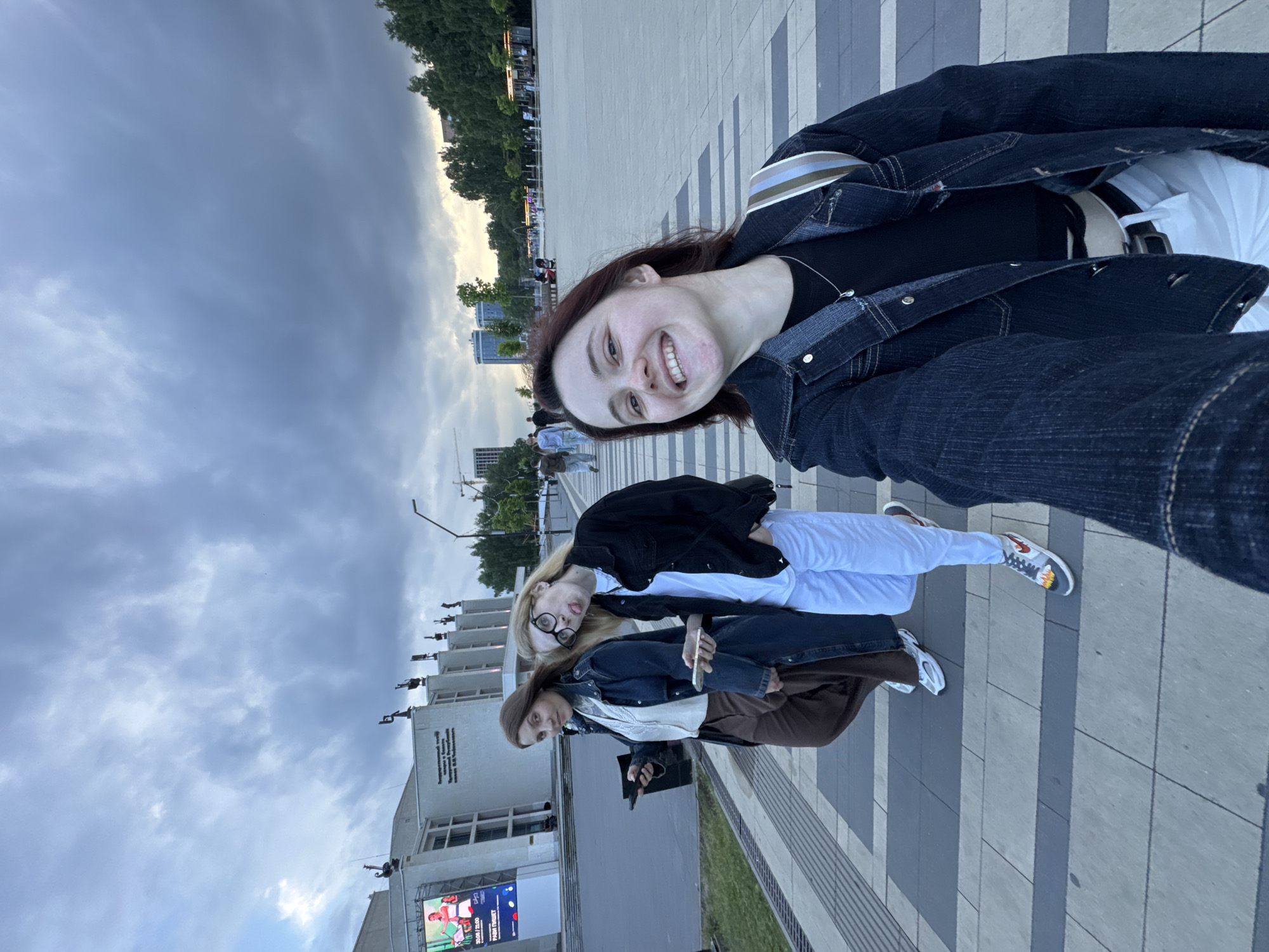 In Izhevsk, Russia, three young women are captured in a selfie style photograph on a paved plaza. The woman closest to the camera, in the foreground, is smiling widely and wearing a dark denim jacket over a black top and white pants, with a striped strap bag slung over her shoulder. Behind her, two other women are walking. The woman in the middle is wearing a black jacket over a white shirt and white pants, sticking her tongue out and wearing glasses. The woman on the far left is wearing a dark trench coat over a white blouse and brown pants. In the background, a large modern building with a marquee displaying an advertisement for an event can be seen, possibly the Udmurt State Opera and Ballet Theatre, given the statues on its roof. The sky above is overcast with dramatic clouds, and trees line the edge of the plaza. The paving stones are arranged in a geometric pattern of light and dark gray. Other people are visible in the distance, suggesting a public space.