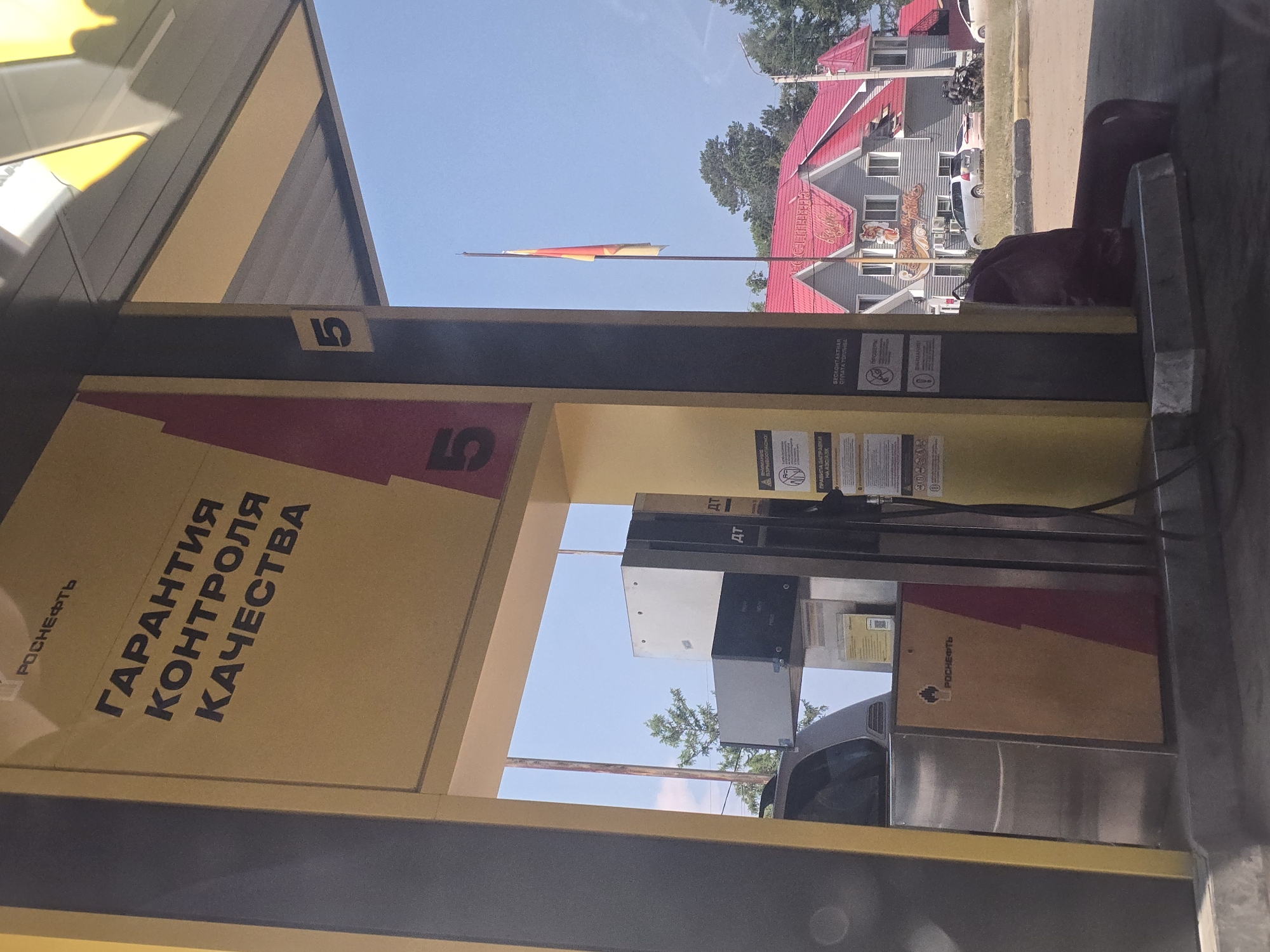 The image depicts a gas station in Turka, Russia. The primary subject is a Rosneft branded fuel pump with a large yellow and red sign on its side displaying "ГАРАНТИЯ КОНТРОЛЯ КАЧЕСТВА" (Guarantee of Quality Control). The number "5" is prominently displayed on the pump structure. To the left of the pump, a silver car is partially visible. In the background, a building with a red roof and the Cyrillic text "РОСПЕЧАТЬ" (Rospechat) is visible. A flag, appearing to be red, yellow, and white, flies from a pole in front of the building. The environment is outdoors under a clear blue sky with some trees visible. The ground surface appears to be asphalt. There are no people clearly visible in the image.
