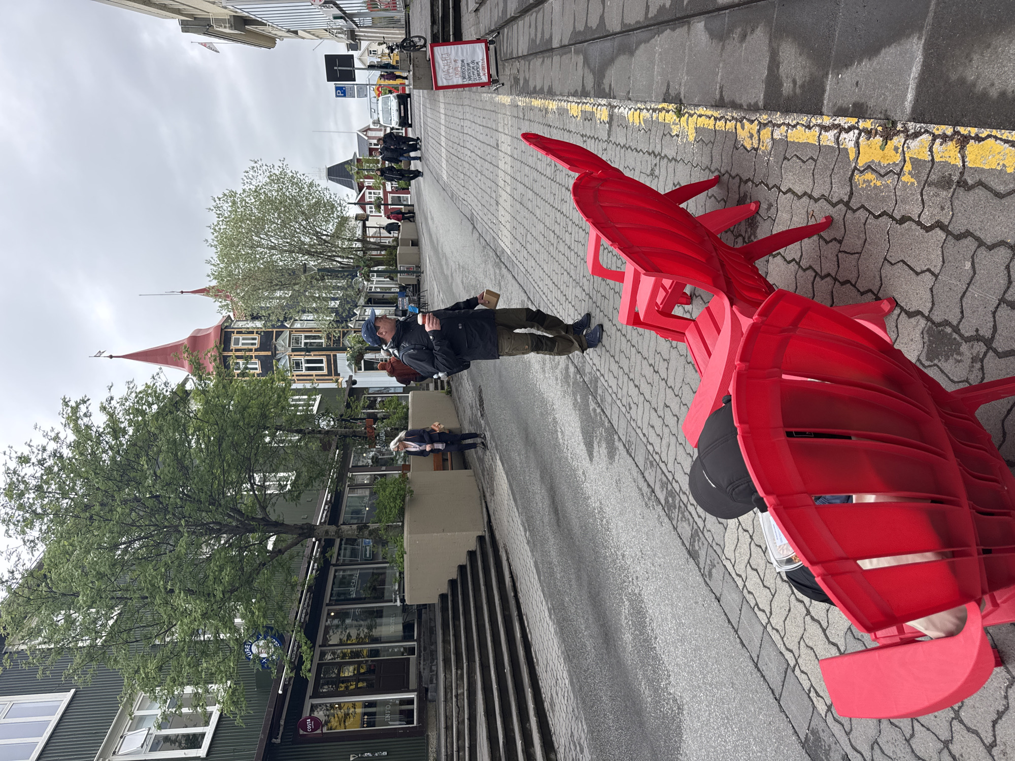 This street-level photograph depicts an urban scene in Akureyri, Iceland, on an overcast day. The image is taken from a slightly elevated perspective, looking down a paved street lined with buildings and scattered pedestrians.
In the immediate foreground, two large, bright red Adirondack-style chairs are positioned on a grey paver sidewalk. One chair is occupied by a person whose upper body is obscured, wearing a black baseball cap. The sidewalk abuts a damp, dark asphalt road, distinguished by a worn yellow line painted along its edge.
A man in a dark jacket, olive-green trousers, and a blue cap walks on the sidewalk, holding a small, light-colored box or cup in one hand and another item in the other. He is also carrying a backpack. Further down the street, several other individuals are visible, suggesting general pedestrian activity.
The buildings lining the street display varied architecture. On the left side, a notable structure features a striking red, pointed roof and dark siding with white-framed windows, characteristic of traditional Nordic style. A verdant tree with light green foliage stands next to it. Other buildings feature corrugated green siding, and one displays a partial blue circular sign with "COSTA" visible, indicating a Costa Coffee. Another sign reads "INFO". In the distance, a red A-frame sign with unreadable white text is present, along with a blue "P" parking sign. The sky is uniformly grey and overcast, implying cool weather conditions. The street appears wet, possibly from recent rain.