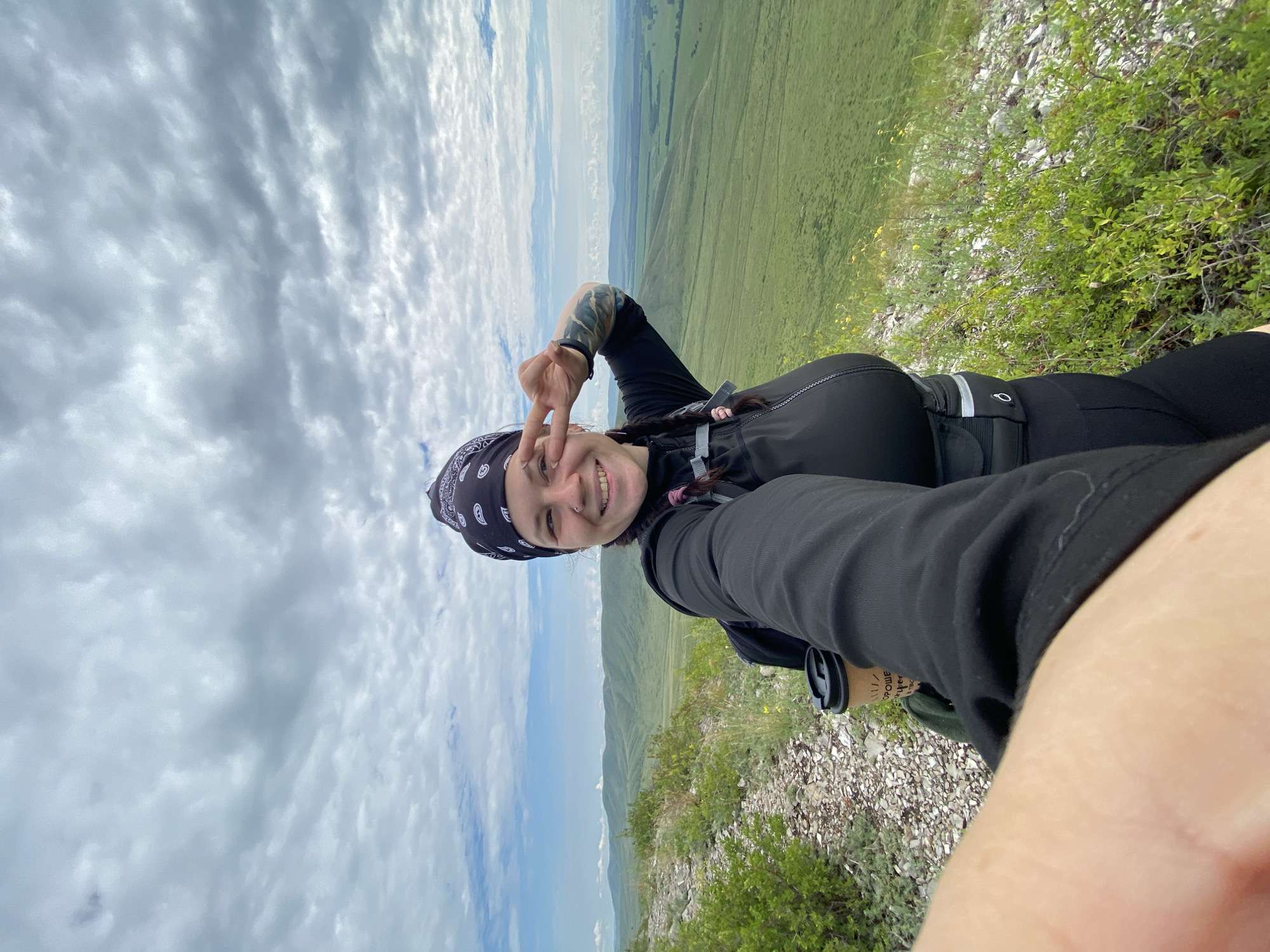 A young woman with dark hair in two braids, wearing a black bandana, black long-sleeved shirt, and black leggings, stands on a rocky, grassy hillside. She holds a disposable coffee cup in her left hand and is making a peace sign with her right hand, partially covering her eye. She is smiling at the camera, taking a selfie. Behind her, a vast, green valley stretches out under a cloudy sky. The landscape appears to be a rural or natural area, possibly a park or nature reserve. The woman is wearing a backpack and a waist pack. The image is taken from a low angle, looking up towards the woman and the sky. The location is identified as Verkhnyaya Chernorechka, Russia.