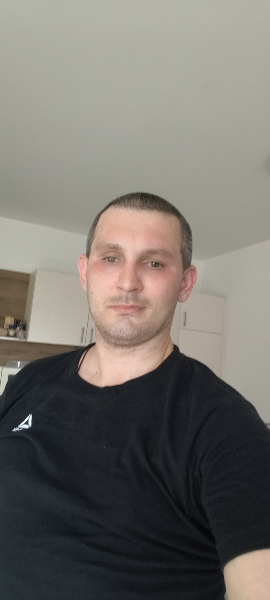 An eye-level, medium shot shows a man with fair skin and short, dark hair, looking directly at the camera. He has light brown eyes and a faint stubble on his chin and upper lip. His cheeks are slightly flushed. He is wearing a black t-shirt with a white Reebok logo on the left side of his chest. The man is indoors, in what appears to be a kitchen or living area. Behind him, in the background, are white cabinets and a wooden countertop. The lighting is bright and diffused, suggesting it's daytime. The image quality is moderately clear.