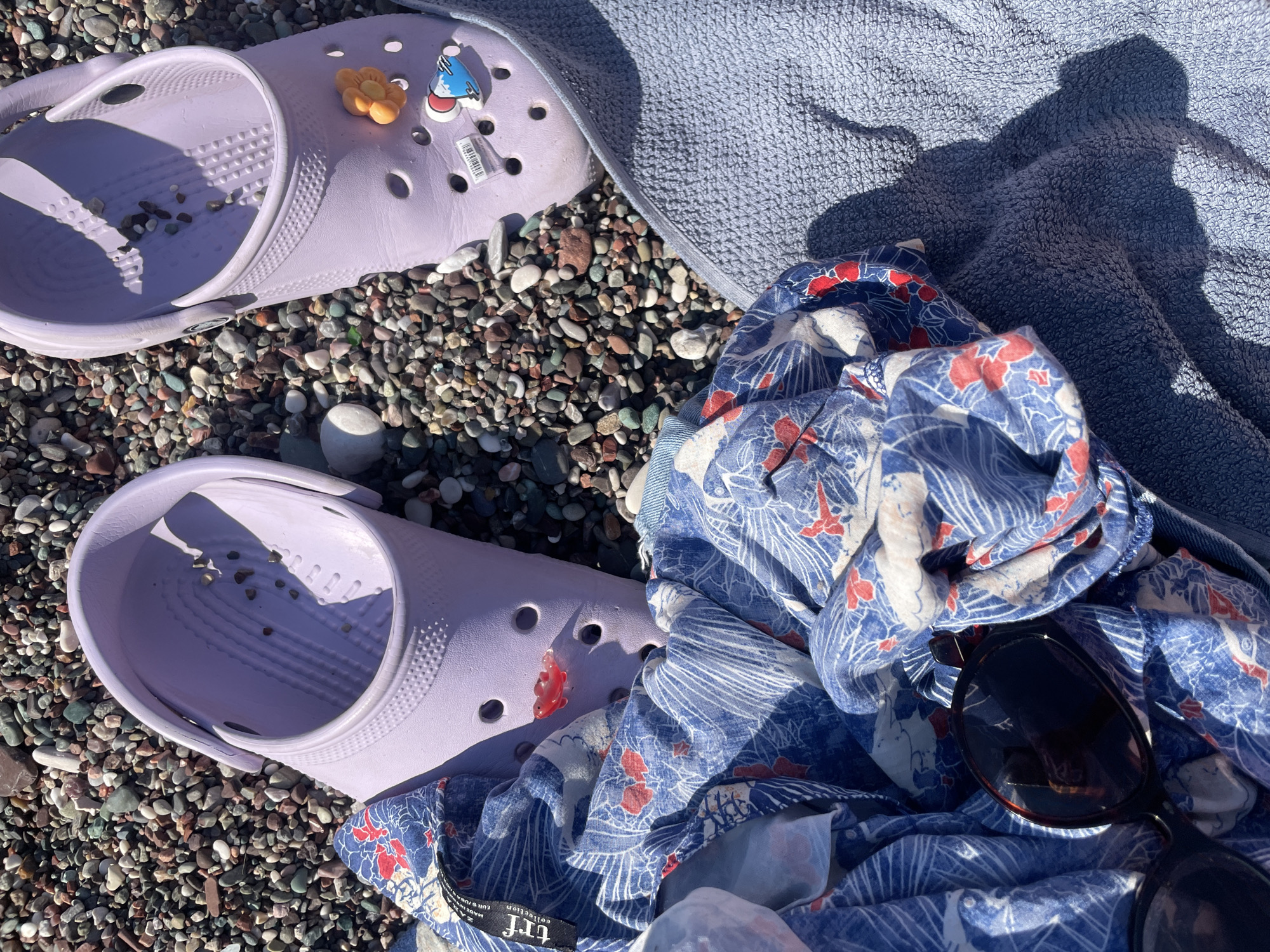 On a pebble beach in Rafailovići, Montenegro, a pair of lavender Crocs sit next to a blue and white patterned fabric. One shoe has a small orange flower charm and a cartoonish charm depicting a person on a mountain. A pair of dark sunglasses rests on the fabric, which is on top of a light blue towel. The scene is bathed in sunlight, casting shadows across the beach and towel.