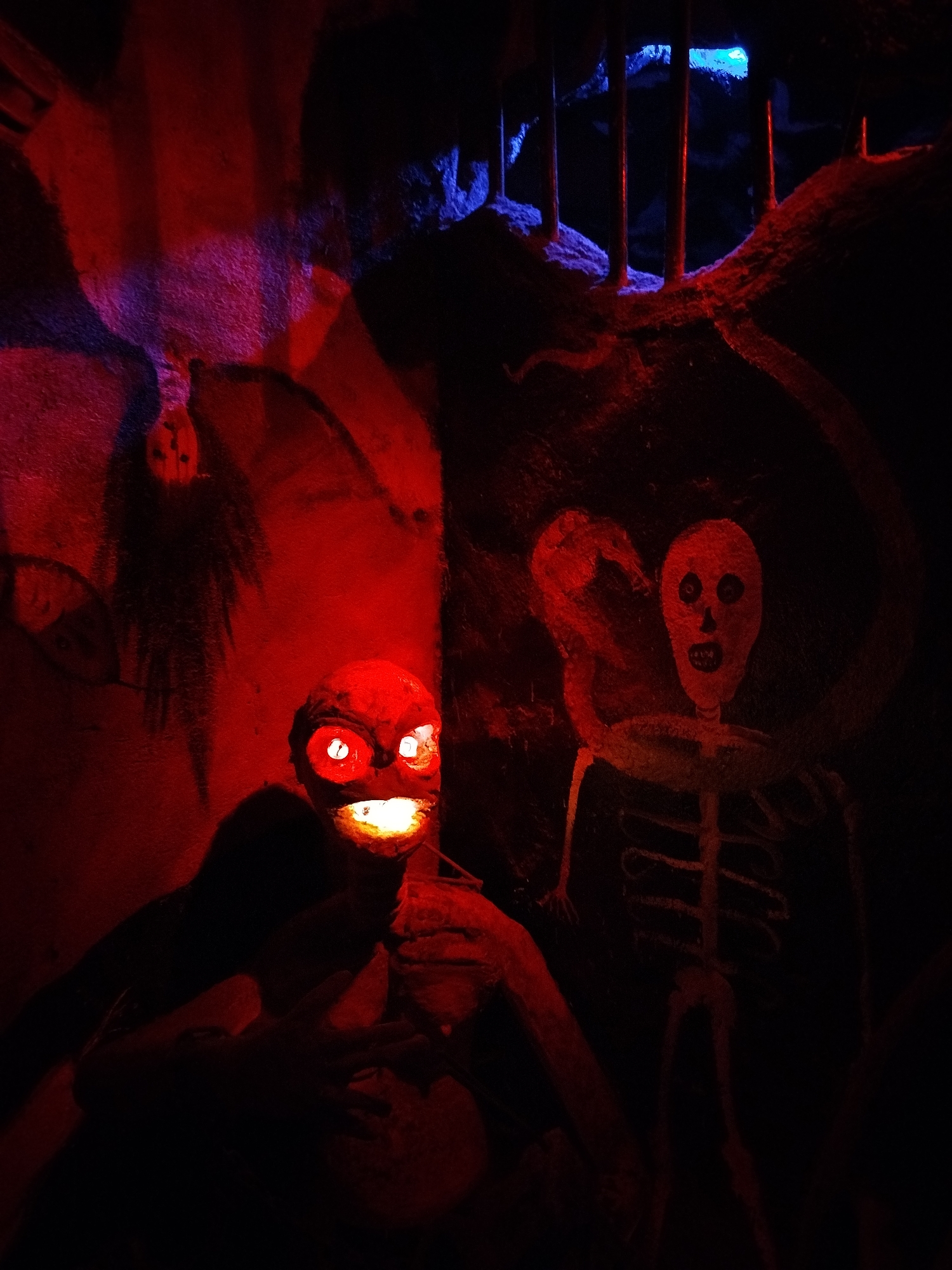 The image depicts a dimly lit scene, possibly in a haunted house attraction located in the city of Unknown, Unknown. In the foreground, a menacing figure with glowing red eyes and a glowing mouth is partially obscured by shadow. The figure appears to be holding something in its hands. Behind the figure, a wall features several spooky elements. An upside-down, ghostly figure is painted on the wall, and to the right, a roughly drawn skeleton with a raised arm is visible. Bars are at the top. The lighting is primarily red, with some blue light near the top of the frame, contributing to the eerie atmosphere.