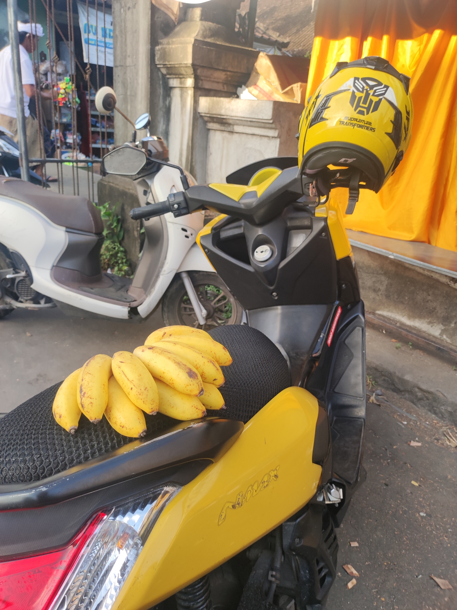 In Ubud District, Indonesia, a yellow and black Yamaha NMAX scooter, with a Transformers-branded yellow helmet hanging from the handlebars, is parked on a paved street. A bunch of bananas sits on the black seat of the scooter. Behind the NMAX, a second white scooter is parked. In the background, a partially visible shop with a man in a white shirt inside can be seen behind a barred window. A golden curtain is seen to the right of the scooter. The street appears to be in a semi-urban area.