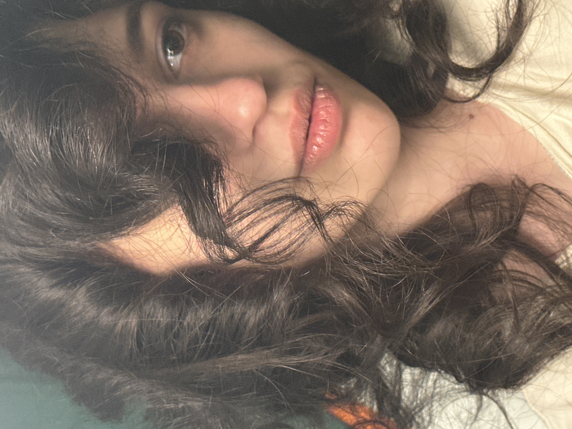 A close-up, head-and-shoulders shot of a young woman with dark, wavy hair framing her face. She is looking slightly to her right, with her hair partially obscuring her left eye and forehead. Her lips are slightly parted and appear to have a glossy finish. The lighting casts a soft glow on her face, highlighting her cheekbone and nose. The background is blurred, with a hint of green on the left and a light-colored garment on her shoulders. The image appears to have been taken indoors.