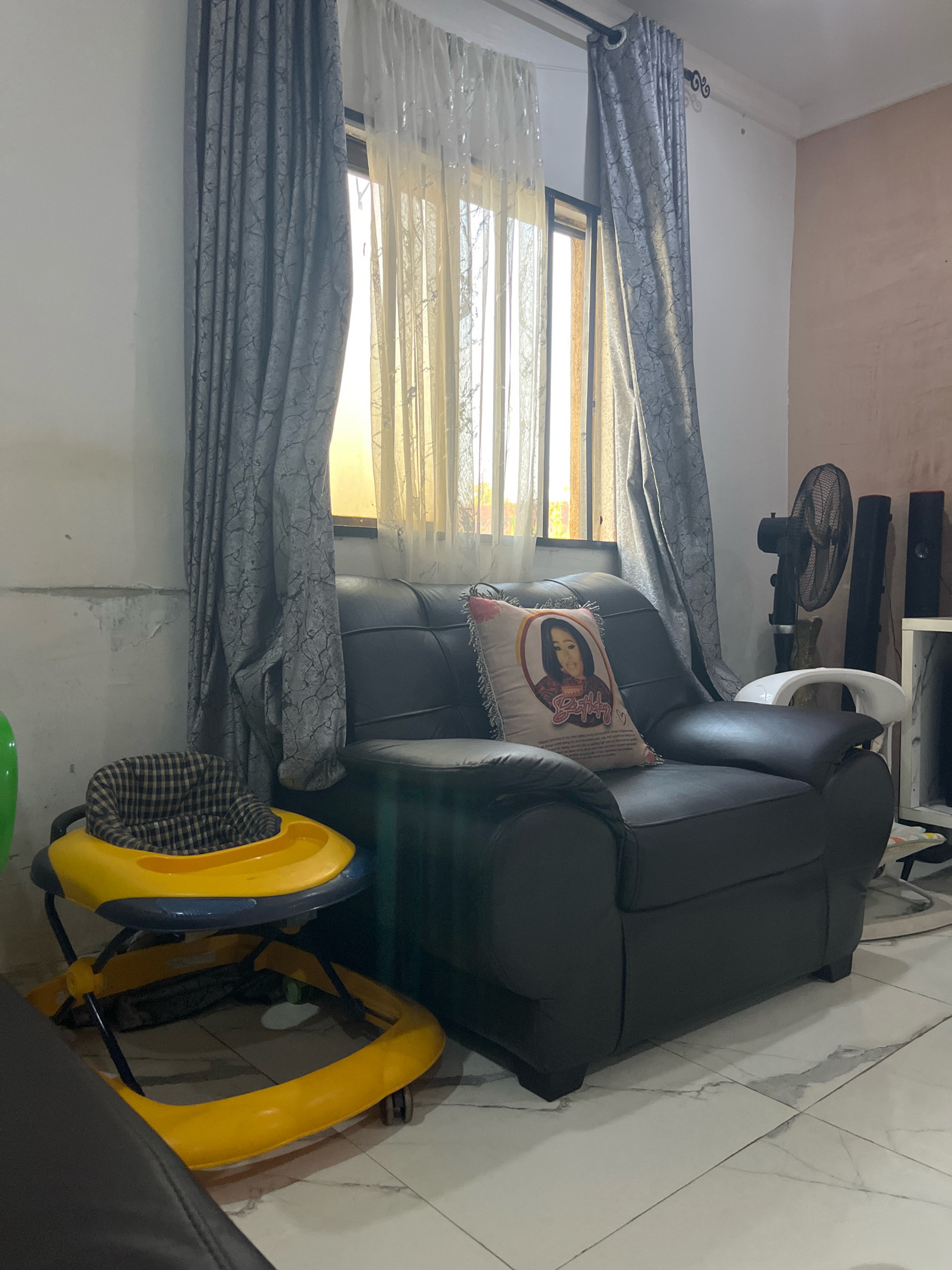 The image depicts an indoor scene, likely a living room, with a focus on furniture and window treatments. A dark brown, leather armchair is prominently placed in the center, adorned with a decorative pillow featuring a woman's portrait and text. To the left of the armchair, a yellow baby walker with a checkered seat insert is visible. The background features a window covered by dual layers of curtains: an outer layer of gray, patterned fabric and an inner layer of sheer, off-white material. Sunlight filters through the sheer curtains, illuminating the space with a warm glow. To the right of the armchair, a black standing fan and two dark cylindrical objects, possibly speakers, are situated next to a white shelving unit. The floor is tiled with a marble-like pattern. The walls appear to be painted a light color, with some signs of wear and discoloration. The scene suggests a domestic setting, potentially in a residential area of Jos, Nigeria, given the implied geographical context.