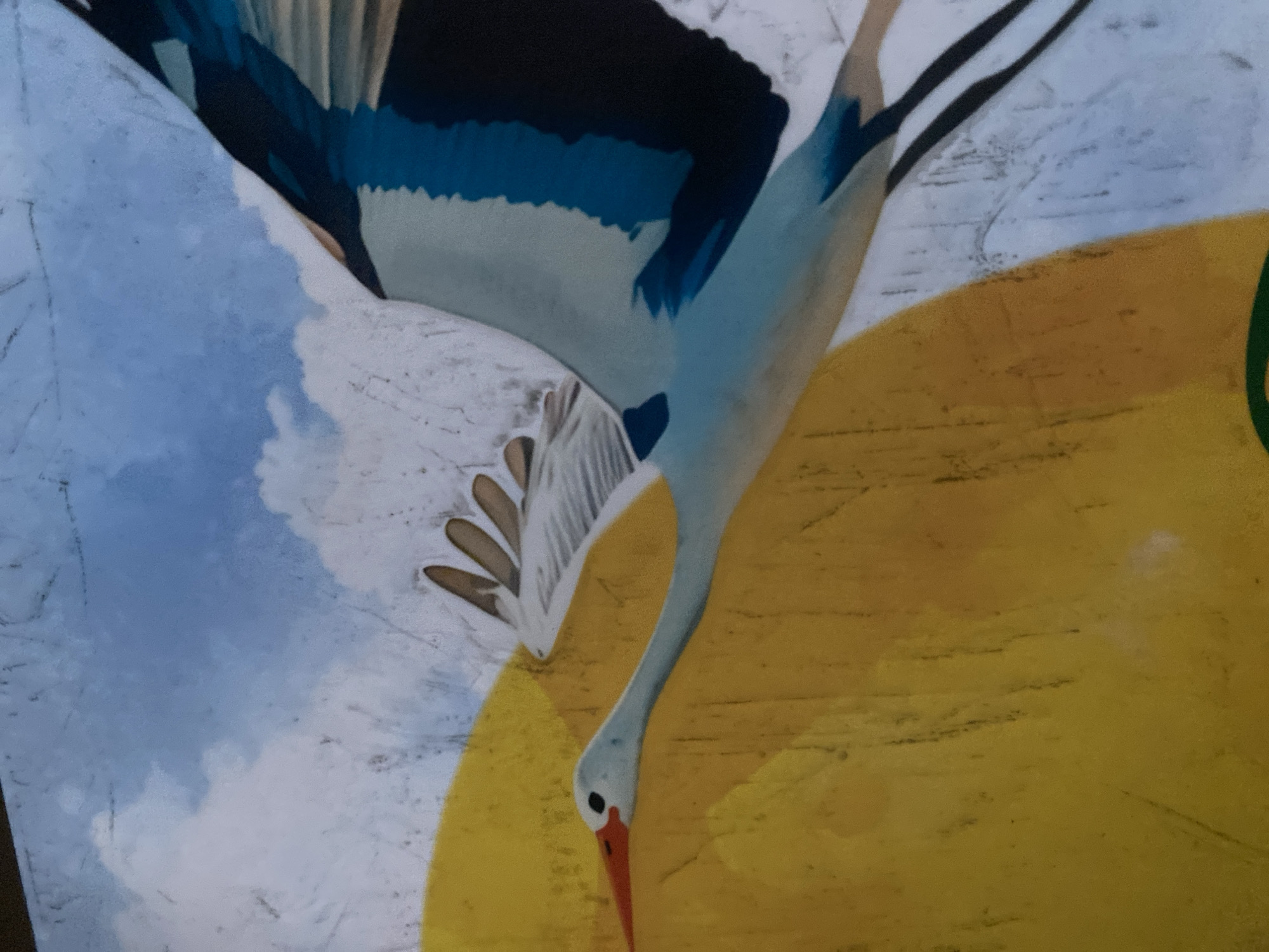 A painting depicts a stork in flight against a backdrop of a bright yellow sun and a blue sky with white clouds. The stork is rendered in profile, with its long, orange beak, black eye, and light blue body. Its wings are spread wide, showing feathers of white, blue, and black. The painting has a textured appearance, possibly due to the medium or surface it is on, with visible brushstrokes or lines. The style is somewhat illustrative, with bold colors and clear outlines. The location of this painting within the city of Unknown, Unknown is not determinable from the image.