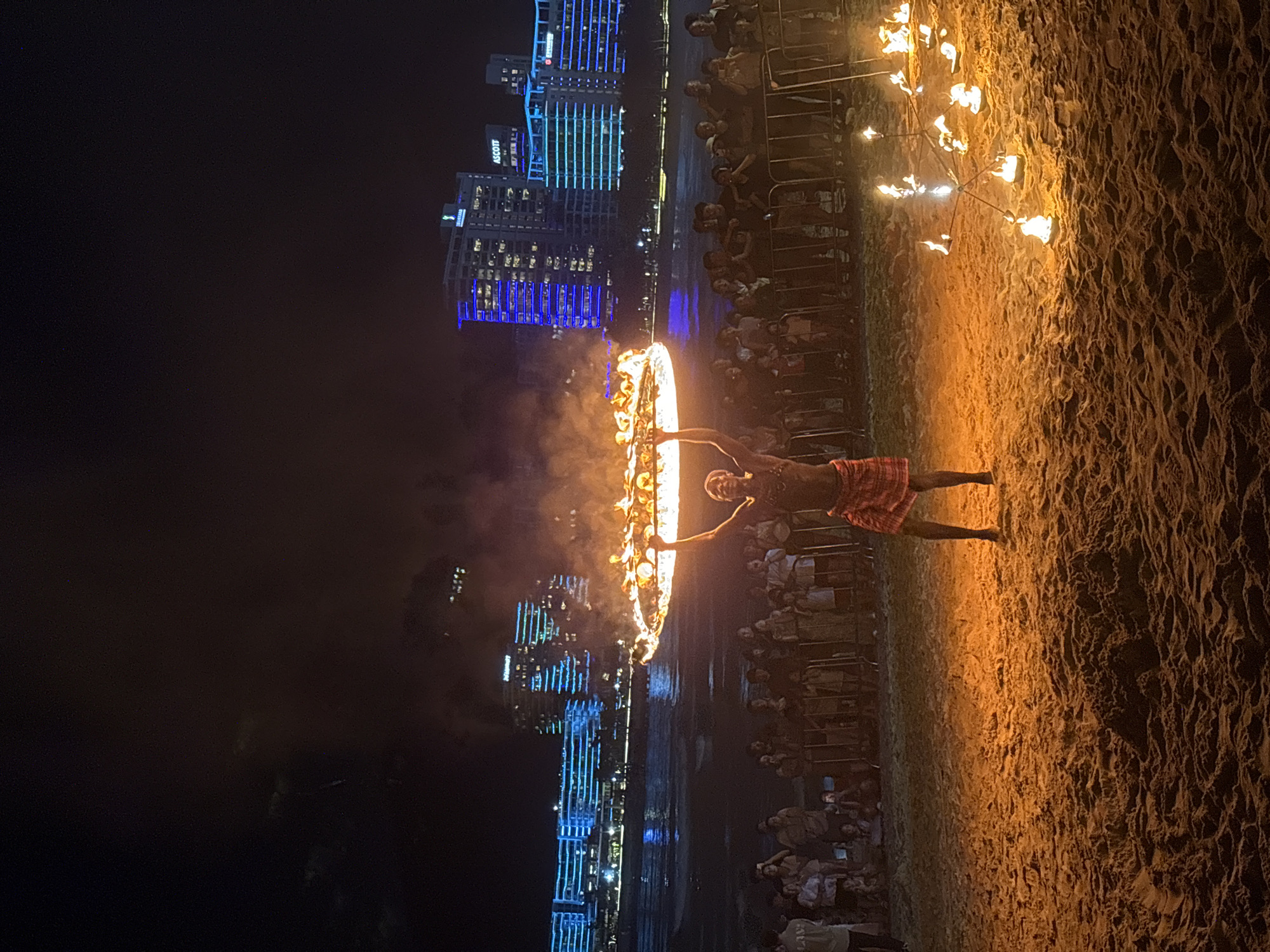This is a nighttime photograph of a fire dance performance on a sandy beach. A performer, wearing a red and white patterned loincloth, stands in the center holding a large flaming hoop above their head. Smoke billows upwards from the hoop. In the foreground, more smaller flaming devices are scattered on the sand. A crowd of spectators stands behind a barrier, watching the performance. In the background, the ocean is visible with gentle waves, and beyond that, several modern buildings are illuminated with blue and green lights. The sky is dark and cloudy. The location is identified as the city of Jiyang District, China.