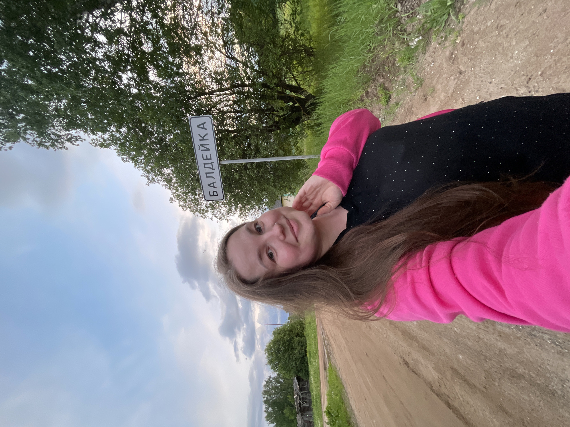 A selfie shows a light-skinned young woman in the foreground, wearing a black top with small white dots and a pink long-sleeved jacket. She has light brown hair. Her right hand is near her face. Behind her is a road sign with the word "БАЛДЕЙКА" written in Cyrillic letters. The background includes a dirt road, trees, and a cloudy sky. Some grassy vegetation borders the road. In the distance, there is some indistinct wooden construction or dilapidated structure. The overall scene suggests the subject is located in the village of Балдейка in Russia.