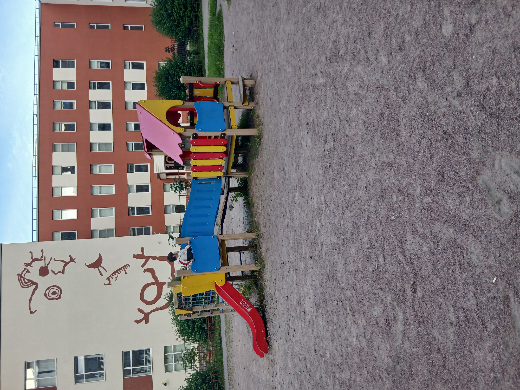 In the city of Murino, Russia, a child is standing on a colorful children's playground structure, which includes a red slide, blue and yellow platforms, and a pink roof. The playground is situated on a gravel surface, adjacent to a building with abstract red mural art on its facade. Another building, predominantly red with white window frames, stands to the right. Green bushes and grass are visible in the background. The scene appears to be outdoors in an urban residential area.