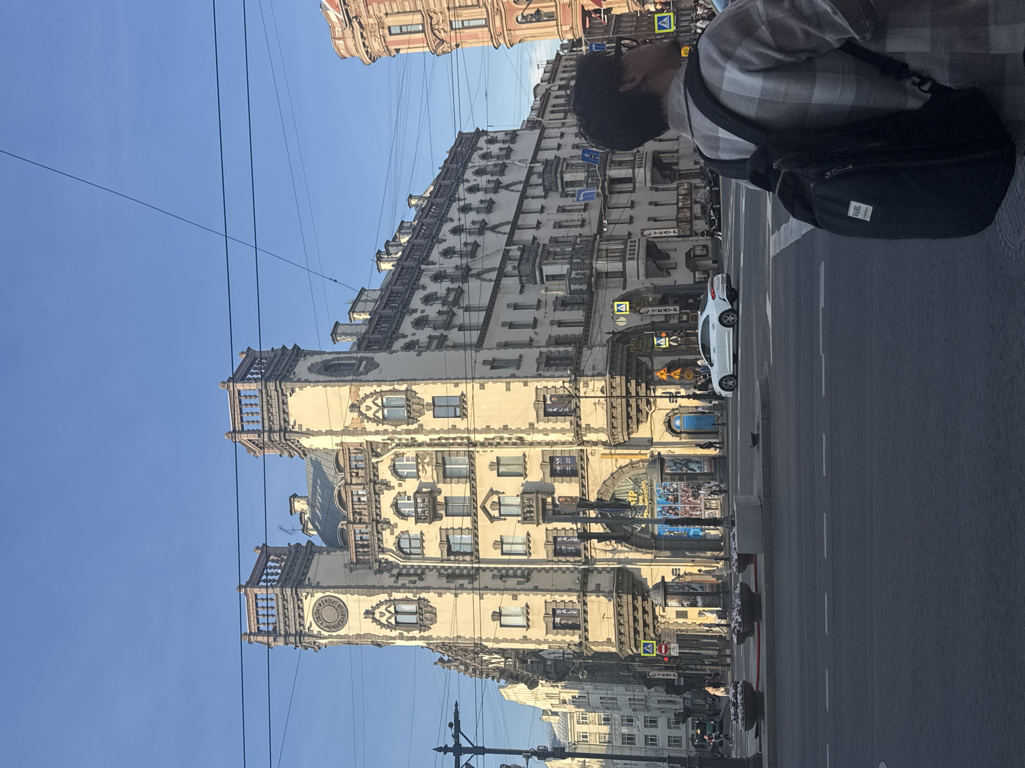 In Saint Petersburg, Russia, an individual wearing a plaid shirt and a backpack stands on a sidewalk, looking away from the camera. Behind them, an ornate cream-colored building with a clock tower and multiple windows dominates the scene. The building features decorative balconies and intricate architectural details. Across the street, a white car is visible, and further down the road, other buildings and pedestrians can be seen. The sky is clear and blue, with overhead wires crisscrossing the frame. The scene appears to be a busy urban street during daylight hours.