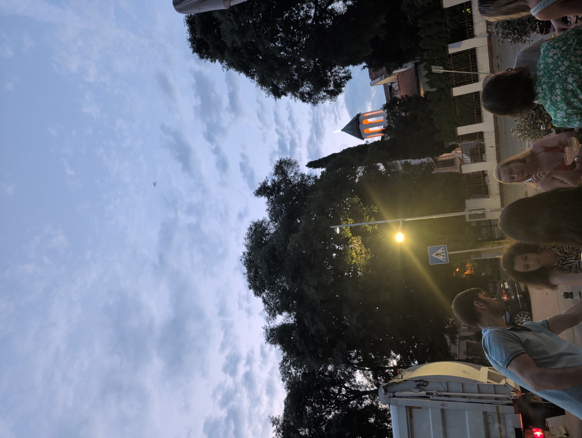The image depicts an outdoor scene at dusk, possibly in Varna, Bulgaria, with a group of people gathered around a table. In the foreground, a man in a light blue polo shirt is visible from the side, his attention directed downwards. To his right, a woman with dark, curly hair wears a patterned top. Further to the right, a blonde woman in a red dress holds a cigarette and a glass. The background is dominated by lush green trees, and a prominent church tower with illuminated windows stands out against the cloudy sky. A garbage truck is parked on the left side of the frame, and a street light and a pedestrian crossing sign are also present. The overall atmosphere suggests a casual gathering in an urban setting as evening approaches.