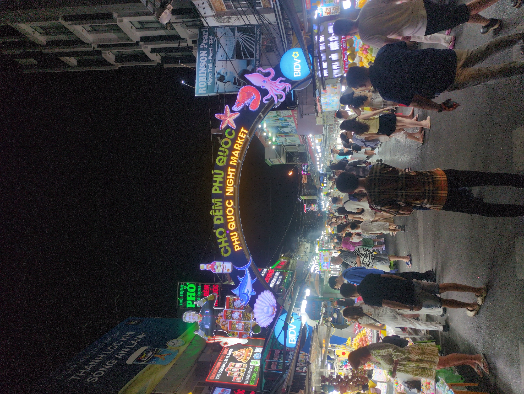 This is a nighttime view of the Phú Quốc Night Market in Vietnam. A large arched sign with sea creature decorations marks the entrance. Below the sign is a densely packed crowd of people walking through the market. Many vendors and illuminated signs line the street. One sign advertises "Robinson Pearl" and another advertises "Chouchou Phú Quốc," with 30 flavors. Further down the street, a sign reads "The Thinh Pho." Streetlights and vendor lights create a bright and busy atmosphere.