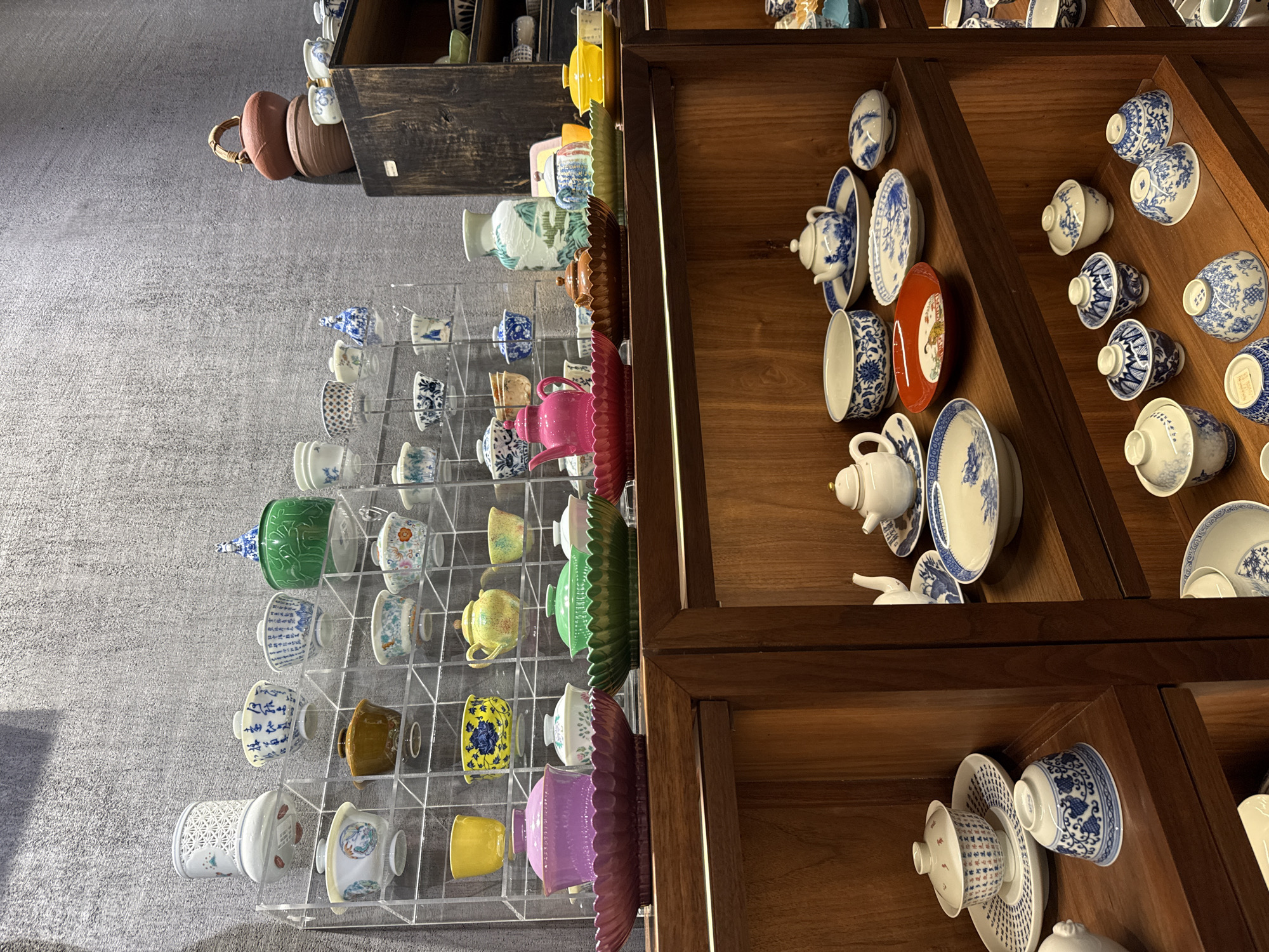 The image depicts shelves displaying porcelain tea sets and decorative items. The shelves are made of dark wood and acrylic. The items range from traditional blue and white porcelain to more colorful pieces. The location is likely in the Liwan District of China, based on the objects presented.