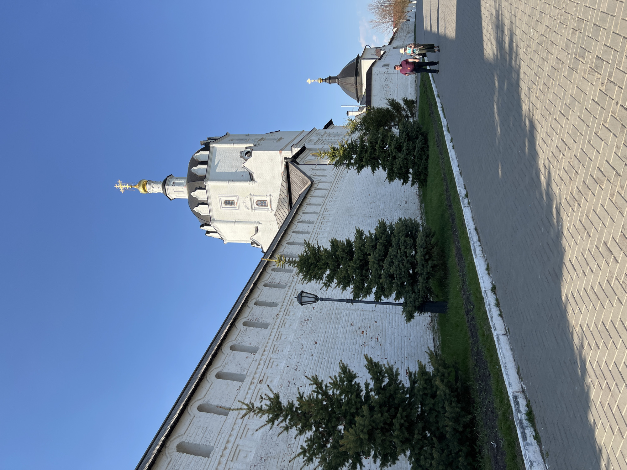 A vertical shot shows the Church of St. Sergius of Radonezh in Sviyazhsk, Russia. The church tower is whitewashed, featuring an ornate golden cross atop a black dome. A thick white wall extends along the side, punctuated by small, arched window-like openings and a black street lamp. Several young evergreen trees line the base of the wall. To the right, a brick walkway extends away, and in the distance, two figures stroll along the path. The sky is a clear, bright blue.