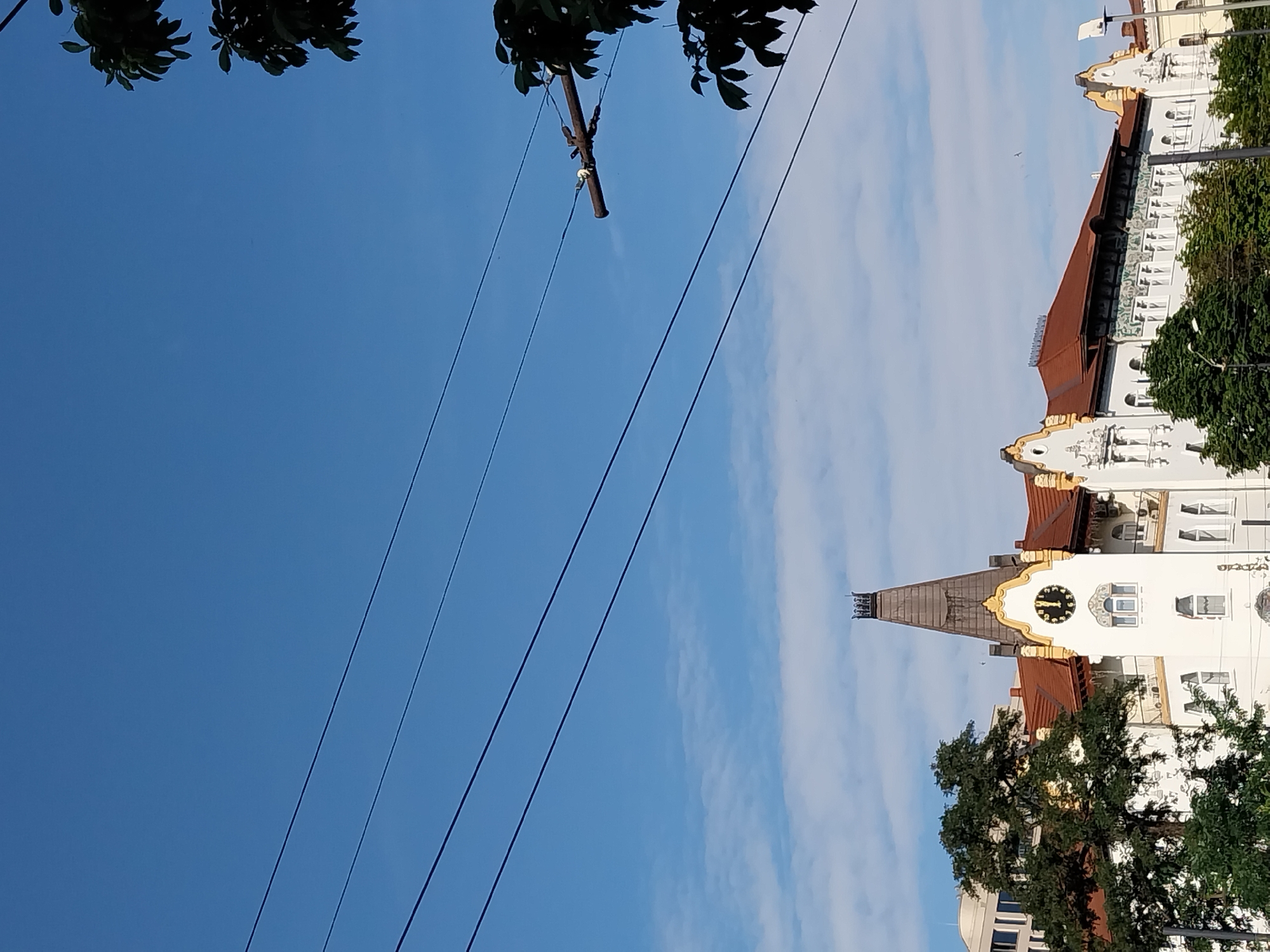 The image captures a view of a building in Dnipro, Ukraine, under a clear blue sky with scattered clouds. The building features a prominent clock tower with a pointed roof, adorned with decorative elements. Several trees are visible in the foreground, partially obscuring the lower portions of the building. Overhead, power lines stretch across the frame, with a wooden pole supporting some of the wires. The architecture suggests an older, possibly historical, structure. The lighting indicates it is daytime, likely late morning or early afternoon, given the brightness and the position of the sun. There are no people visible in the image.