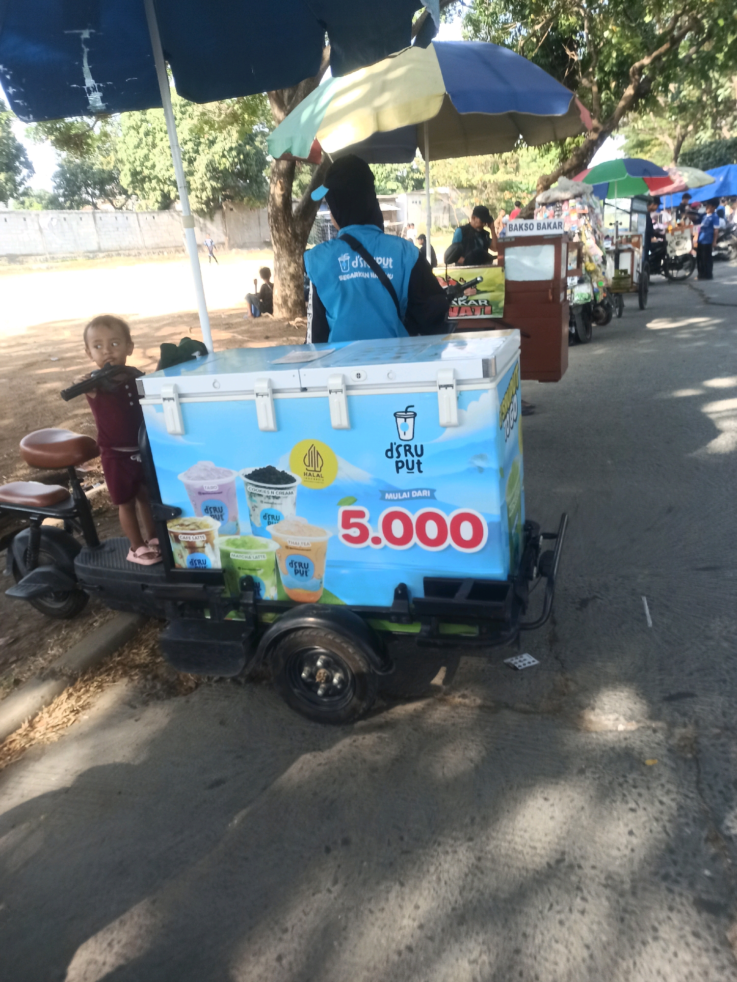 In Telagamurni, Indonesia, a young child stands next to a mobile beverage cart under an umbrella. The cart is blue and features images of various drinks like taro, cookies and cream, cafe latte, matcha latte, and Thai tea. A price of 5,000 is prominently displayed. A person wearing a blue vest stands behind the cart, facing away from the camera, with the logo "DS/RU PUT" visible on their back. In the background, other vendors with carts are visible, and the area appears to be outdoors with trees and a concrete wall. The ground is asphalt, and shadows suggest it is a sunny day.