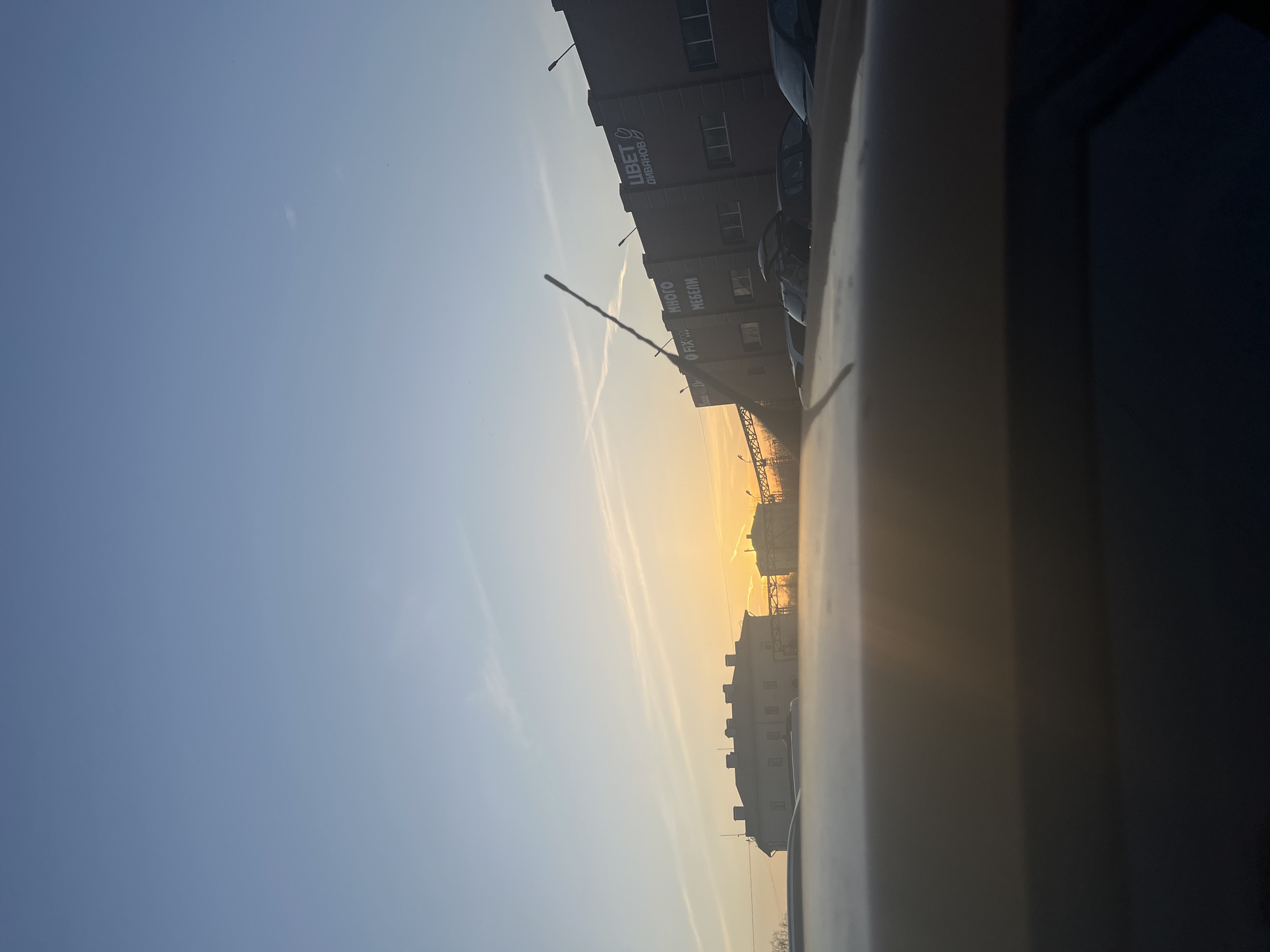 The image depicts an urban landscape in Klin, Russia, captured from the interior of a vehicle, looking towards a low sun on the horizon. The sky transitions from a clear light blue overhead to bright orange and yellow directly above the horizon line, indicative of either sunrise or sunset. Several contrails are visible in the sky, forming an "X" pattern in the upper left and a linear trail extending from the sun.
In the midground on the left, a light-colored, multi-story building with a dark roof and multiple windows is visible. To the right, a dark, modern commercial building with vertical architectural paneling features several illuminated signs. These signs include "ЦВЕТ ДИВАНОВ" (TSVET DIVANOV - "COLOR OF SOFAS"), "МНОГО МЕБЕЛИ" (MNOGO MEBELI - "LOTS OF FURNITURE"), "FIX PRICE", and a partially visible "БАНК" (BANK) sign. A row of dark-colored passenger vehicles is parked in front of this commercial building; one vehicle has its trunk open. Further in the background, to the right of the central axis, industrial structures featuring a large metal framework or gantry crane are silhouetted against the bright horizon.
The foreground is dominated by the dark, reflective surface of what appears to be the vehicle's dashboard and hood, obscuring the ground level immediately in front of the camera. A thin, dark, vertical object, possibly an antenna or a piece of debris, extends upwards from the dashboard near the center-right of the lower frame.