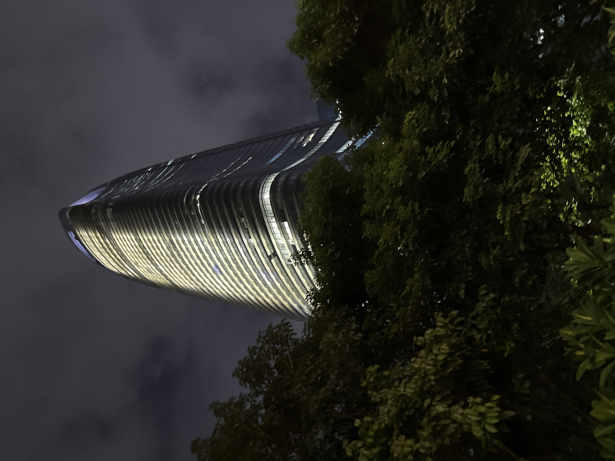 The image captures a modern skyscraper at night, its curved facade illuminated by warm, horizontal lights that accentuate its architectural design. The building appears to be made of glass and metal, with a sleek, futuristic aesthetic. The sky above is dark and cloudy, suggesting it's nighttime or a heavily overcast evening. To the right of the frame, lush green trees with dense foliage partially obscure the view of the skyscraper, creating a contrast between the natural and the man-made. The leaves of the trees are also faintly illuminated, possibly by the building's lights or ambient city light, giving them a slightly luminous quality. The perspective is from below, looking upwards at the towering structure and the overhanging trees, which lends a sense of scale and grandeur to the scene. The overall mood is somewhat dramatic, with the play of light and shadow, the imposing architecture, and the darkness of the night. The location context provided is Haizhu District, China, which places this scene within a likely urban environment. There is no visible text in the image.