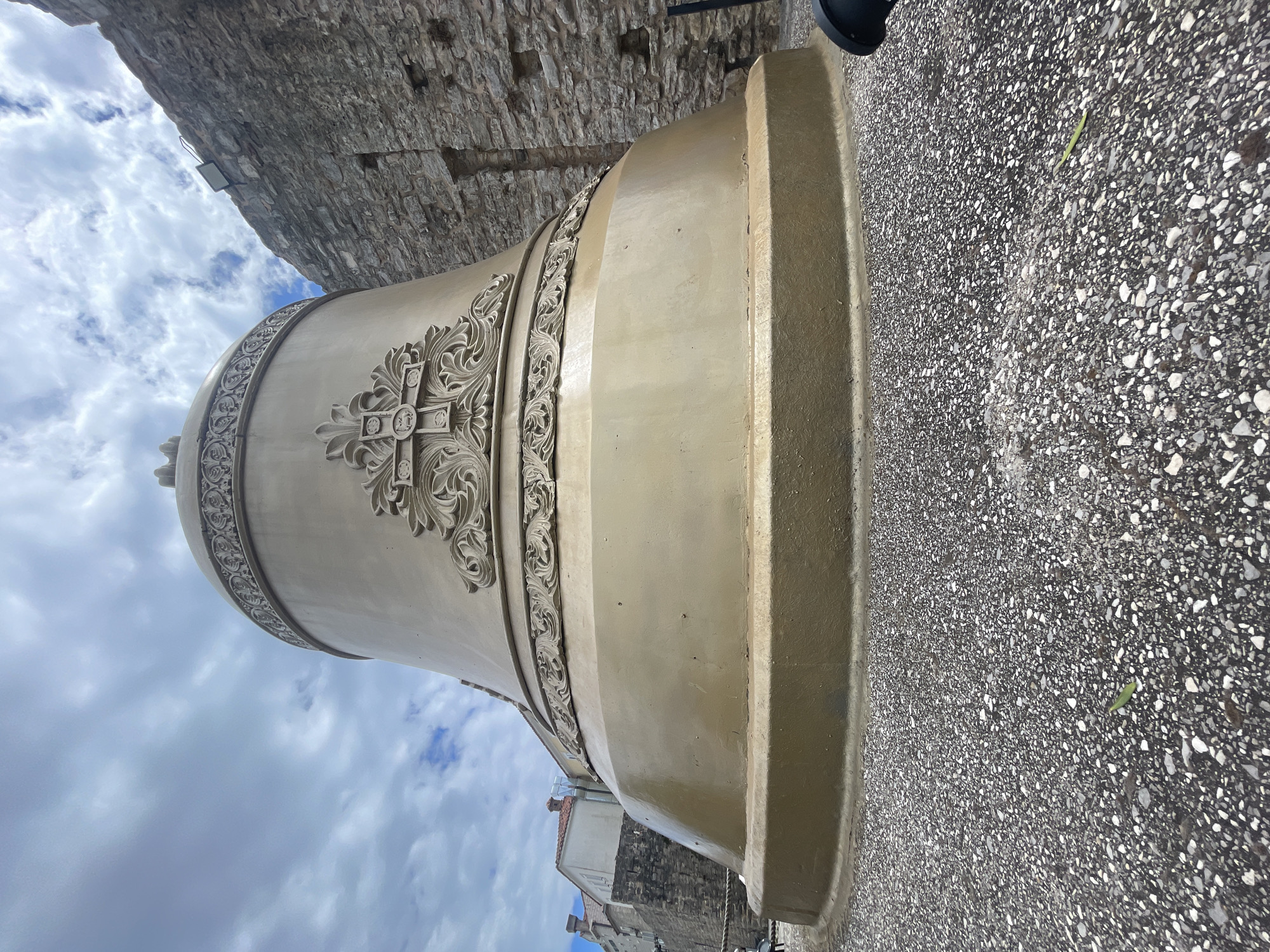 A large, ornate, light-colored bell-shaped object dominates the foreground, viewed from a low angle. The object features elaborate, raised decorative carvings, including a central cross design surrounded by intricate swirling floral or vine motifs. Two bands with similar patterns encircle the upper part of the bell. The ground beneath the bell is a textured surface composed of small, dark and light aggregates, resembling exposed aggregate concrete or gravel, with a single small green leaf visible.
In the background, a tall, rugged stone wall with visible mortar lines rises upwards, suggesting an ancient structure like a fortress or castle wall. A small rectangular floodlight is mounted on this stone wall. Above the wall and the bell, the sky is partly cloudy with patches of blue visible. In the distance, beyond the bell's base, other buildings with terracotta-colored rooftops are partially visible, hinting at an urban or historical setting. A black object, possibly part of a stand or a light fixture, is partially visible at the upper right. The scene is static, captured during the daytime under bright, cloudy conditions. There is no visible text.