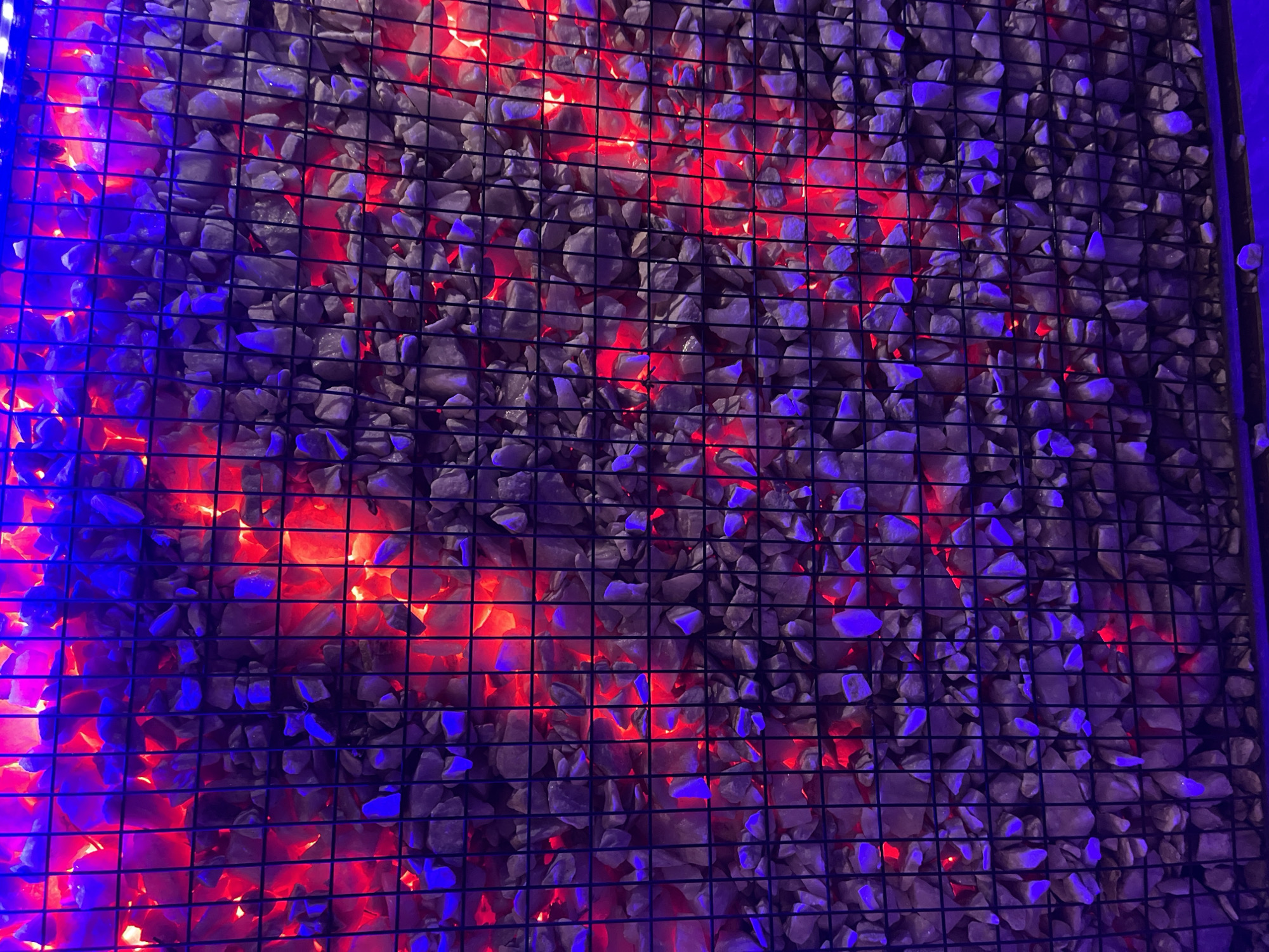 The image shows a wall covered in small, grey stones held in place by a dark wire grid. Red and blue lights illuminate the stones from behind, creating a contrasting color effect. The location in Jos, Nigeria is not evident from the image. There are no people visible in the picture.