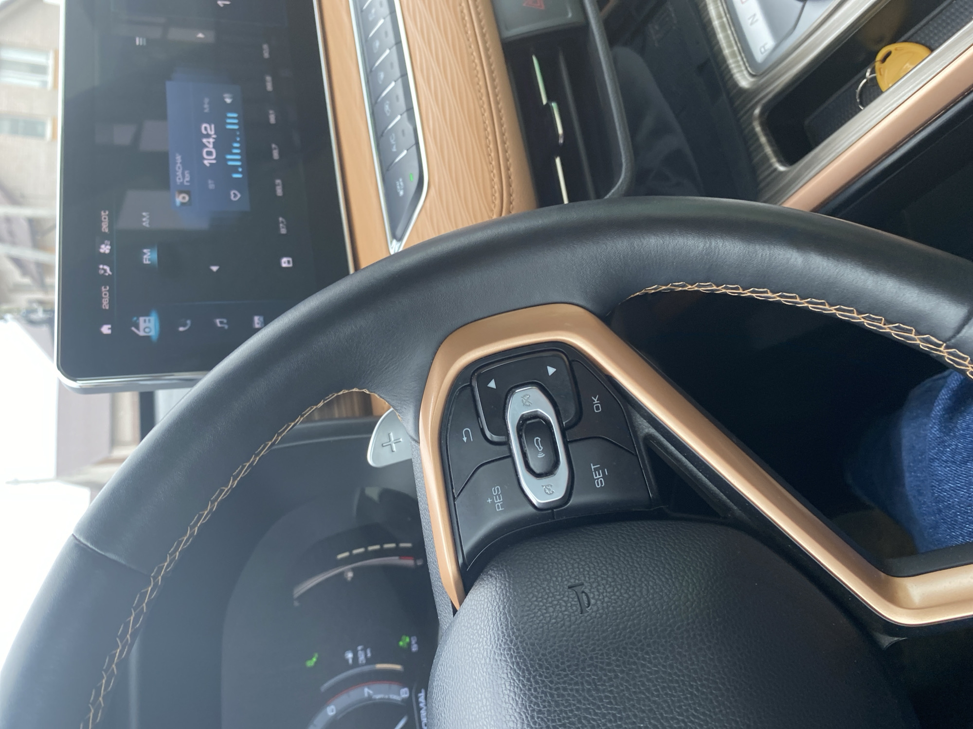 Here's a forensic description of the image:
**Scene:** Interior of a vehicle, specifically focusing on the driver's side.
**Objects:**
* Steering wheel: Predominantly black with a gold accent trim, and visible stitching along the rim. Contains integrated buttons for vehicle controls, including cruise control ("SET," "RES") and menu navigation.
* Dashboard: Includes a central infotainment screen displaying audio information, including a radio station tuned to 104.2 MHz. The climate control settings are also visible (26.0°C). Trim is light brown/beige.
* Gearshift console: partially visible; includes a car key.
* Paddle shifters: Present behind the steering wheel.
**Environment:** A residential area is discernible through the windshield, with buildings visible outside.
**People:** A person's leg, wearing a blue jean can be seen.
**Actions:** No actions are explicitly visible in the image.