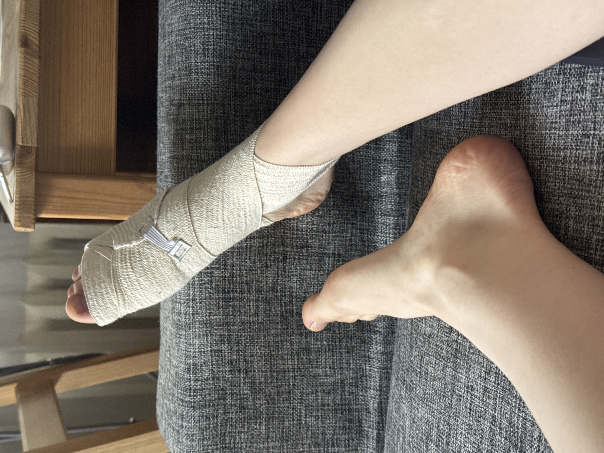 The image shows a close-up, top-down view of a person's lower legs and feet, resting on a grey, textured fabric surface, likely a sofa or bed. One foot is bare, revealing the sole and heel, while the other foot is extensively wrapped in a light-colored, beige elastic bandage. The bandage covers the foot and ankle, secured by a small silver-colored clip or buckle. The toes of the bandaged foot are visible at the end of the wrap.
In the background, to the left, there is wooden furniture with a light brown, natural wood grain finish, suggesting part of a bed frame, chair, or table. The texture of the grey fabric is prominent, appearing to be a woven material. The lighting is soft and even, typical of an indoor setting, without harsh shadows, making it difficult to determine the exact time of day.
The primary activity depicted is resting, with the bandaged foot indicating a possible injury, sprain, or a need for support/recovery. There are no visible emotions or explicit textual elements in the image. The scene conveys a sense of calm and recuperation within an indoor environment, located in Moscow, Russia.