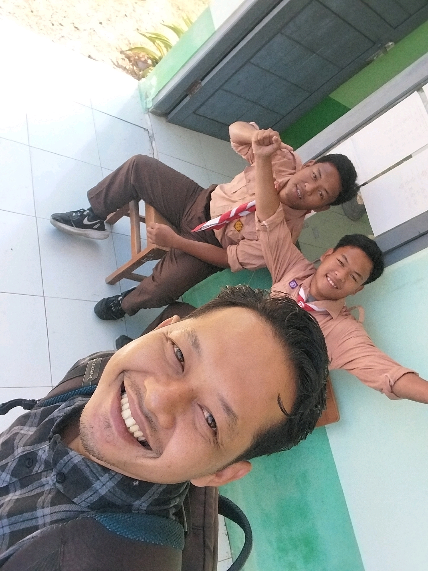 The image, taken in Pandean, Indonesia, shows three young males. The photographer, in the foreground, is looking at the camera and smiling, with his face partially visible. He is wearing a plaid shirt and a backpack. Behind him, two other young males are seated. One is leaning back and giving a thumbs-up, wearing a tan uniform shirt with a red and white scarf. The other is lying down and smiling, also wearing a similar tan uniform shirt and scarf. The background shows a tiled floor and a green wall.