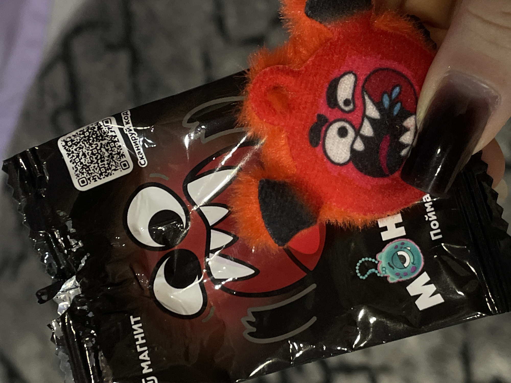 A close-up shot shows a crinkled, black packaging bearing a cartoonish red monster face with large white teeth and circular eyes. A QR code is visible on the upper left of the packaging. The word "МАГНИТ" appears partially visible on the bottom left. Adjacent to the cartoon monster is a red, fuzzy toy that mirrors the face on the packaging. The packaging also features the word "HOW" in white letters along with a teal monster illustration attached to a chain. A human finger with a dark, gradient nail polish holds the package. The location is Chaikovsky, Russia.