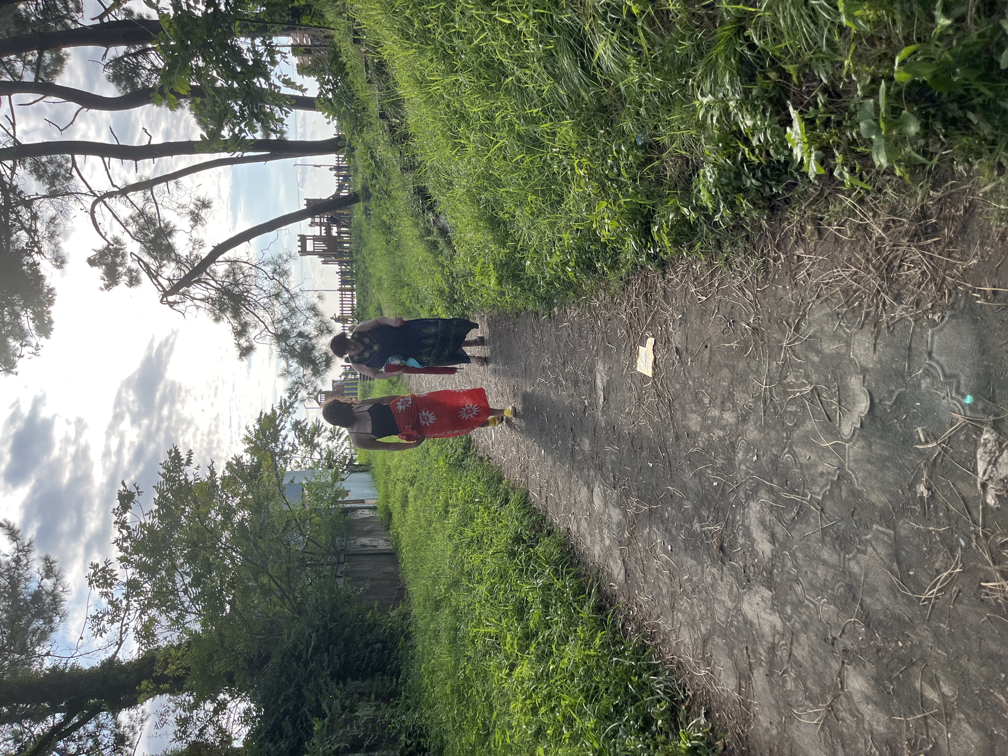 Two individuals, a younger woman in a black top and a red skirt with a sun pattern, and an older woman in a dark patterned dress, are walking away from the camera on a dirt path in Ureki, Georgia. The path is lined with lush green grass and vegetation on either side. In the background, there are trees, a wooden fence, and a playground structure. The sky is partly cloudy, with the sun shining through, casting a soft light on the scene. The overall atmosphere appears relaxed and natural.