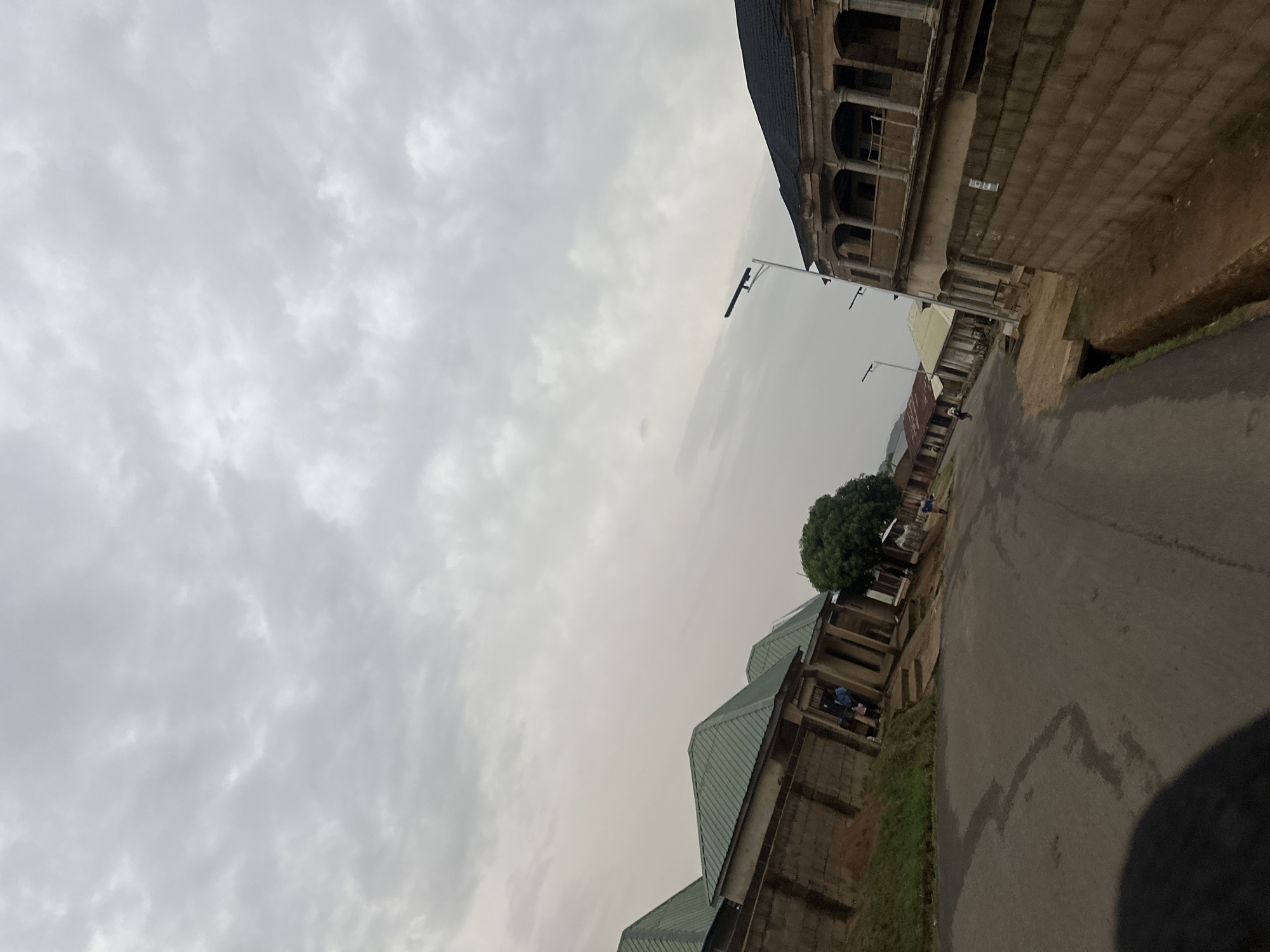 The image depicts a street scene in Jos, Nigeria, under a cloudy sky. A paved road stretches along the right side of the frame, showing signs of recent moisture. Buildings line the road on both sides. The building on the upper right exhibits an older architectural style, possibly with arched openings. Further down the road, more modern structures with green roofs are visible. Street lamps are present along the road. A person is visible walking down the road. A tree stands near the buildings with the green roofs.