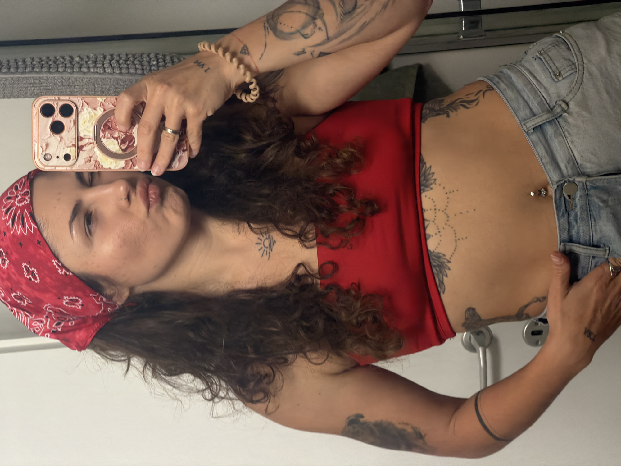 A woman is taking a selfie in what appears to be a bathroom. She is wearing a red bandana, a red top, and light-wash denim shorts. She has multiple tattoos on her arms, chest, and stomach, including a sun, a geometric design, and other various images. She has a belly button piercing. The phone she is using has a floral case. The background shows a white wall with a door handle and a shelf. There is no specific indication of Kiryat Bialik, Israel, or any other city.