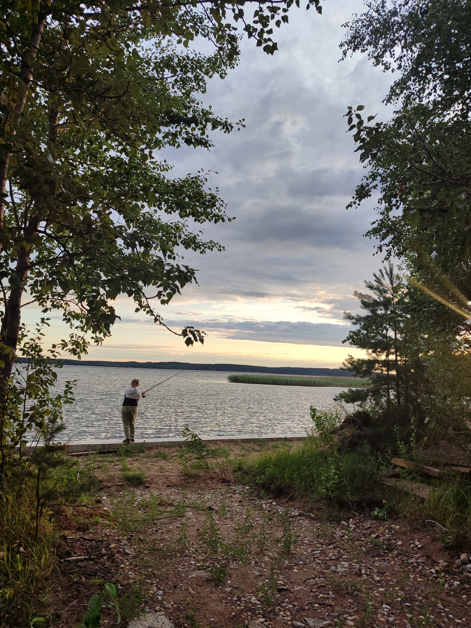 A person is fishing on the shore of a body of water. The person is standing with their back to the camera, holding a fishing rod. The water is calm, with a slight ripple on the surface. The sky is cloudy, with patches of blue showing through. Trees frame the scene on either side, with their leaves rustling in the breeze. The ground is covered with sand and small stones. The sun is setting, casting a warm glow over the landscape. The overall mood of the image is peaceful and serene.