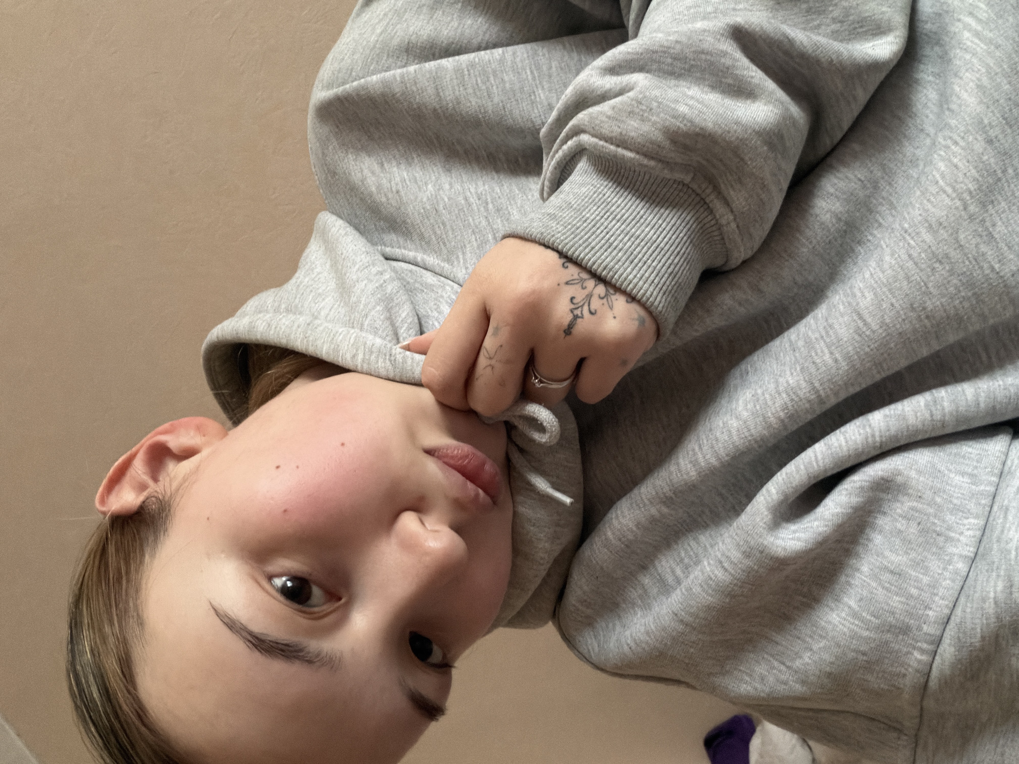 A close-up, tilted selfie shows a person with fair skin, rosy cheeks, and light brown hair wearing a gray hoodie. The person's right hand, adorned with a ring and tattoos, holds the drawstring of the hoodie near their mouth. The background is a plain, light beige wall. A glimpse of a purple and white object appears in the lower right corner, partially obscured. The location is unspecified, and there is no visible evidence to determine if the image was taken in Chaikovsky, Russia.