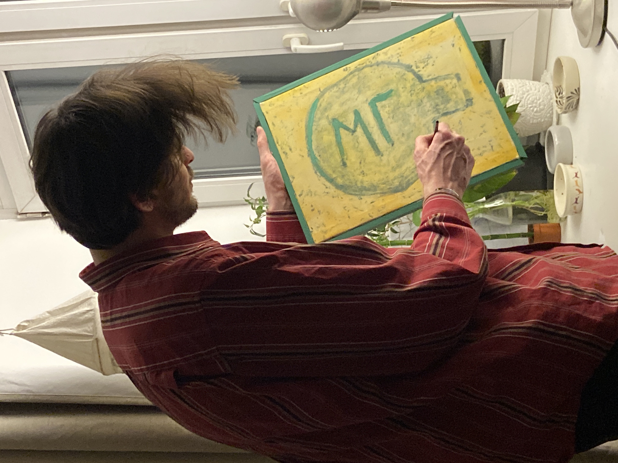 A person, seen from the back, is holding a canvas while drawing on it with a pen. The canvas has a yellow background with green lettering that reads "MF" within a circle, and a geometric shape above it. The person is wearing a red striped shirt. The background includes a window, a lamp, and several small pots with plants. There is no information to suggest that the image was taken in the city of Moscow, Russia.