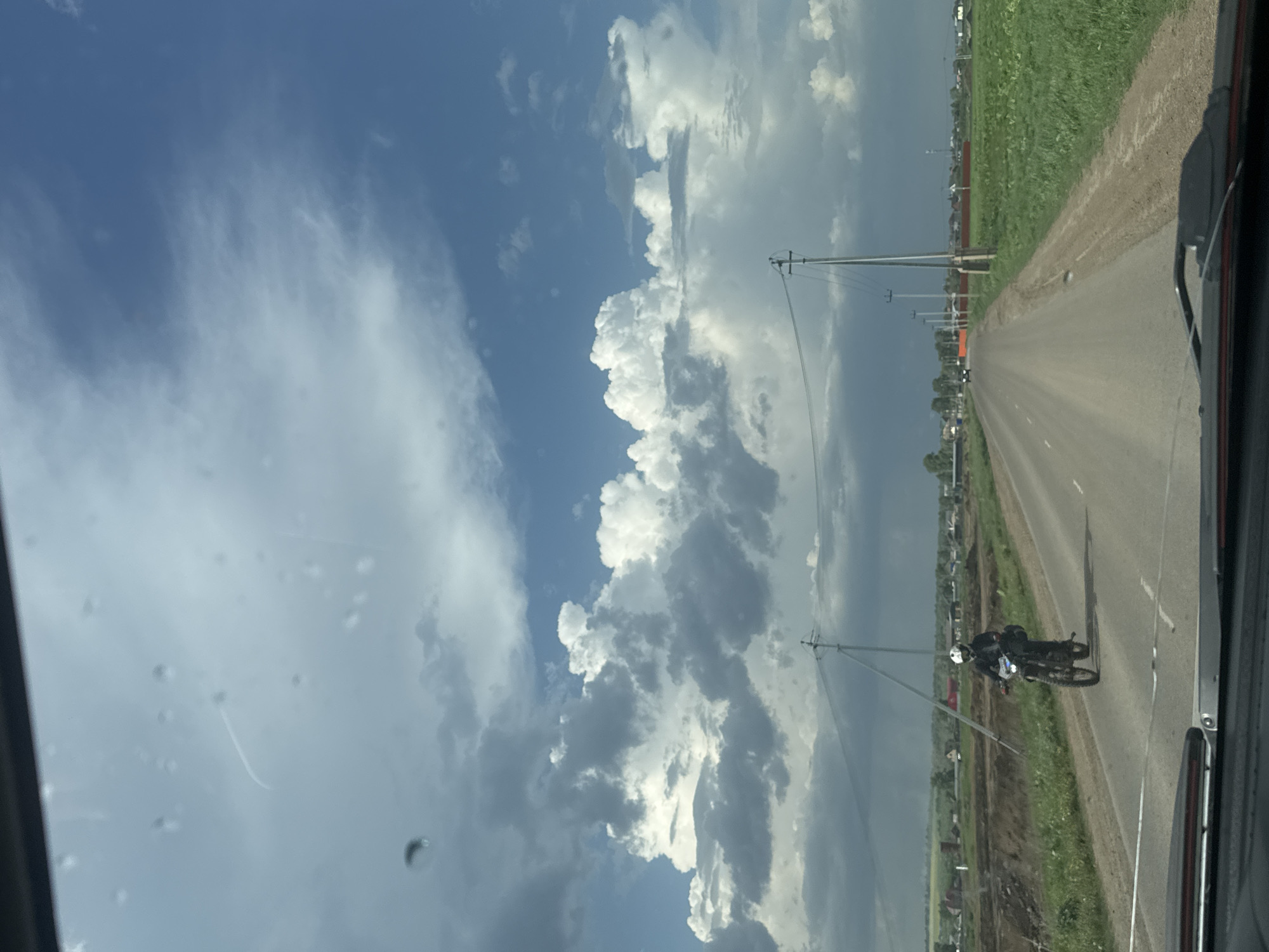 The image shows a view from inside a car, looking down a road in Kitsjanovo, Russia. A motorcyclist is traveling on the road in the same direction as the car. The sky is visible, with significant cloud cover. There are power poles running alongside the road. Fields or grassy areas border the road on both sides. The scene appears to be a rural or semi-rural environment.
