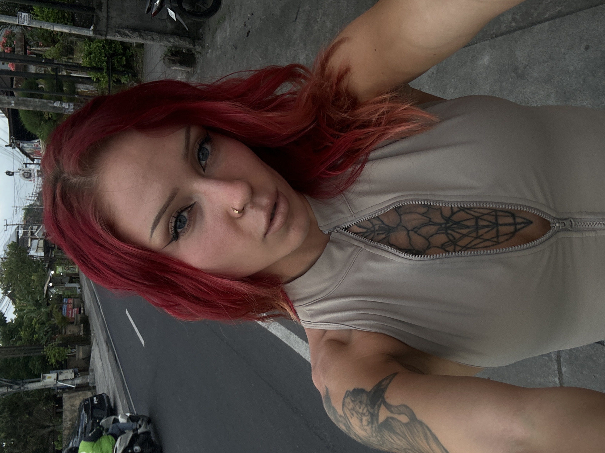 A woman with red hair and light eyes is taking a selfie on a street in North Kuta, Indonesia. She wears a light-colored top with a zipper that is partially open, revealing a tattoo on her chest. Additional tattoos are visible on her arm. The background includes a road with motorcycles parked along the side, a sidewalk, and various buildings and greenery. The lighting suggests it's daytime.