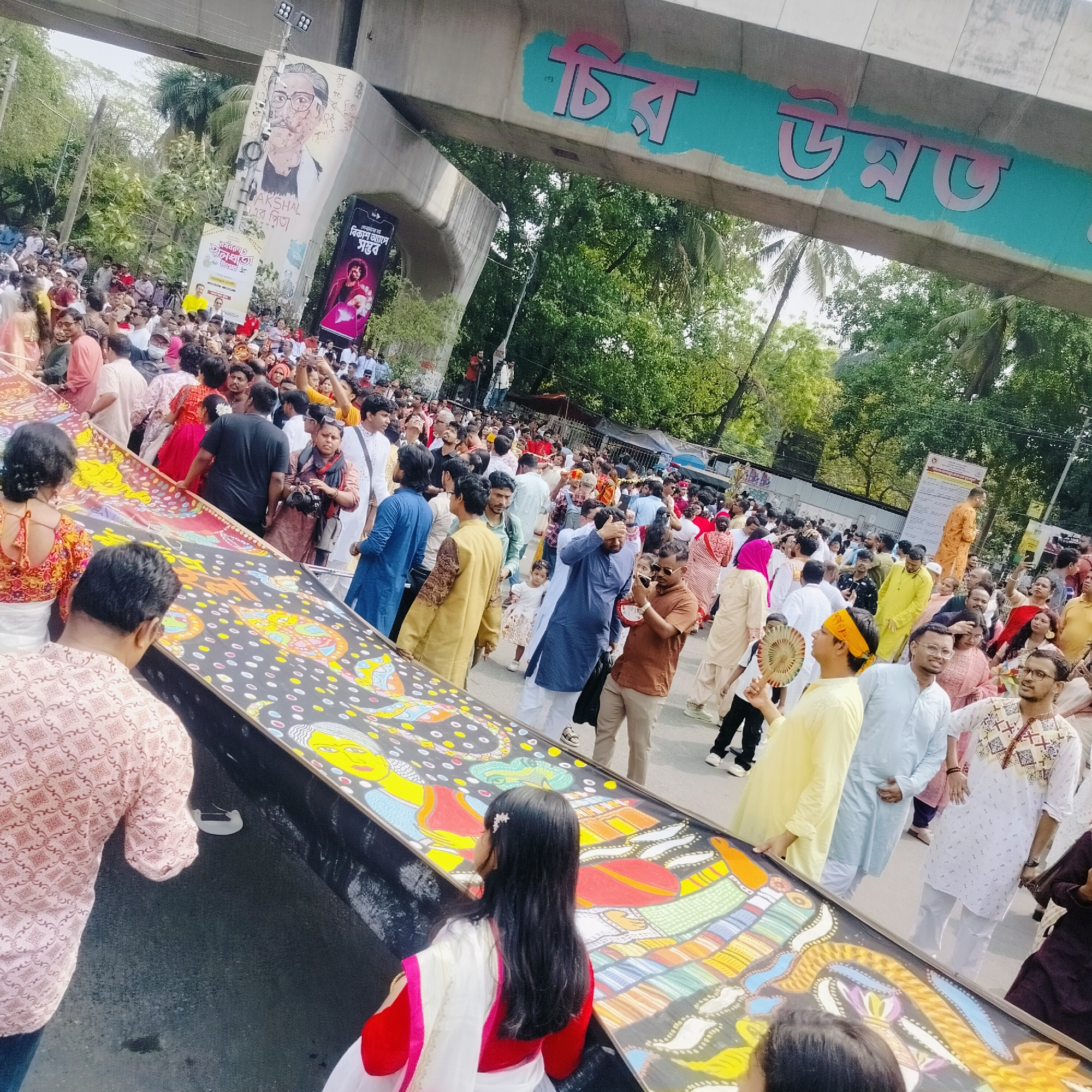 A large crowd of people walk in a street in Dhaka, Bangladesh. They are participating in a procession, carrying a long, painted banner with stylized figures on it. Most people are wearing traditional clothing. Many people are taking photographs. Overhead, a bridge has the Bengali words "Chiro Unmoto" painted on it. A portrait of Sheikh Mujibur Rahman is also visible.