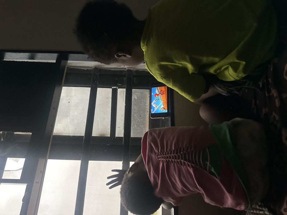 Two children focus on a smartphone screen mounted between window bars. The child in the lime green shirt looks intently at the game, while the other child, wearing a pink shirt, raises a hand slightly. Visible through the window is bright daylight. The children's proximity to the screen suggests a shared focus and perhaps a joint gaming experience.
