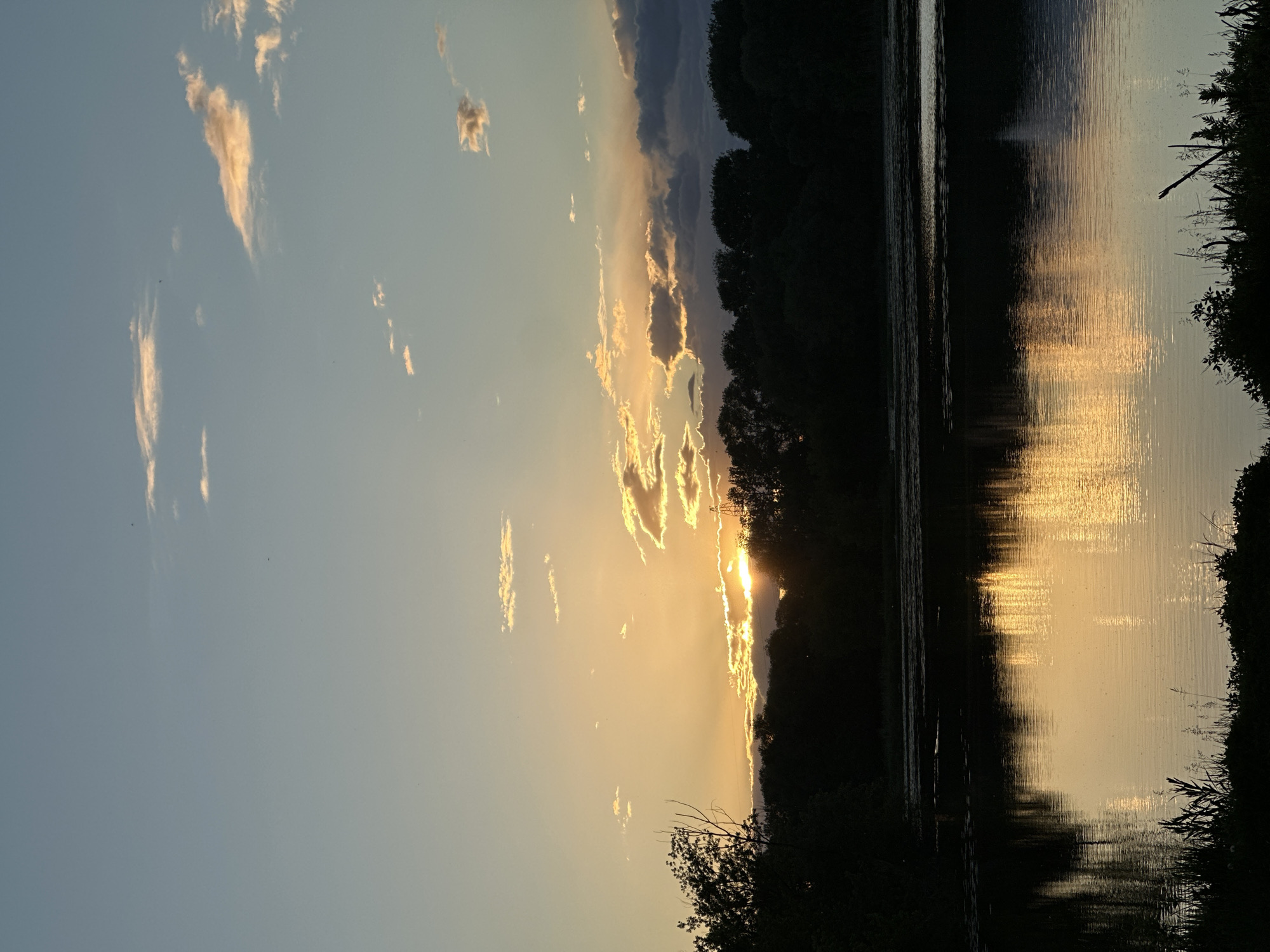 The image depicts a sunset reflected in water, likely a lake or river, in an unspecified location. The sky shows a gradient from light blue to orange-yellow where the sun is setting. Clouds are scattered across the sky, some illuminated by the setting sun. The sun's rays peek through a line of dark trees on the horizon. The water surface reflects the colors of the sky and the light of the sun. Dark vegetation lines the edges of the water. The overall scene conveys a sense of peace and tranquility.