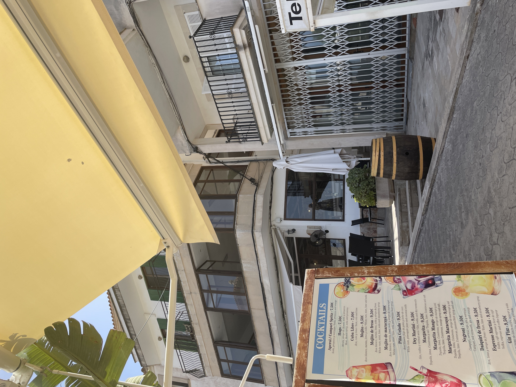 The image shows an outdoor seating area of a restaurant or bar in Santanyí, Spain. A yellow awning extends over a seating area, providing shade. In the foreground, a wooden sign displays a cocktail menu with colorful illustrations of drinks. The menu lists various cocktails with their prices in Euros. Behind the sign, a dark wooden barrel with yellow bands stands on the cobblestone ground. Further back, there are tables and chairs set up, and a building with multiple floors and balconies is visible. A large, rolled-up white umbrella leans against the building. The overall scene suggests a relaxed, sunny atmosphere suitable for outdoor dining or drinks.