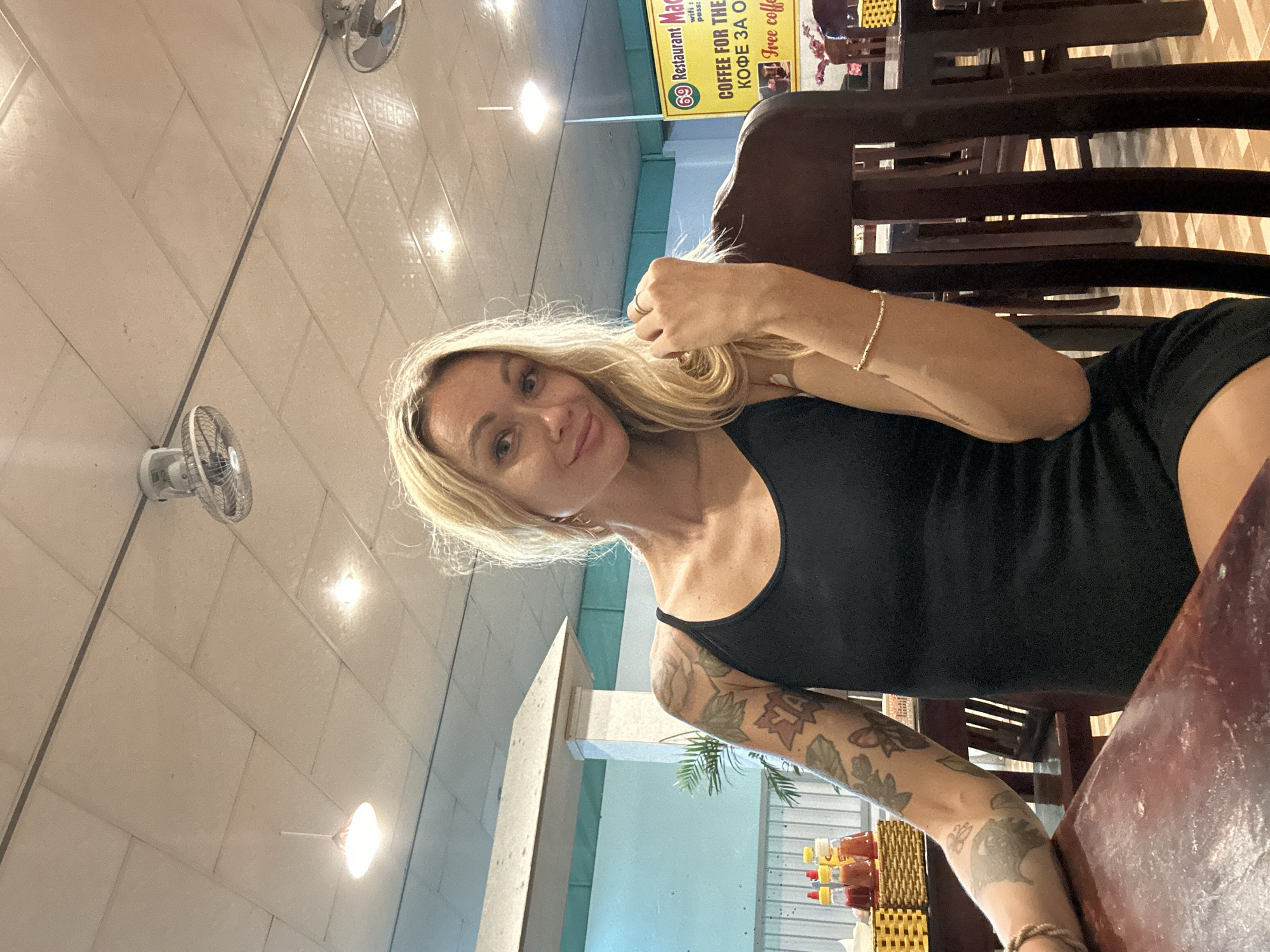 In this vertical, indoor shot taken in Phan Thiết, Vietnam, a woman with blonde hair is seated at a wooden table. She is wearing a black tank top and has tattoos on her arm. Her facial expression is friendly and she appears to be looking towards the camera. Behind her, a yellow sign with text in English and possibly Russian is visible. The sign reads "69 Restaurant Mac" and mentions "COFFEE FOR THE" and "Free coffe". The setting appears to be a casual dining establishment, possibly a cafe or restaurant, with a tiled ceiling and wall. There are fans mounted on the wall, suggesting a warm climate. Wooden chairs and tables are visible in the background, along with some bottles of condiments on another table. The lighting is bright and artificial, indicating it could be daytime or evening.