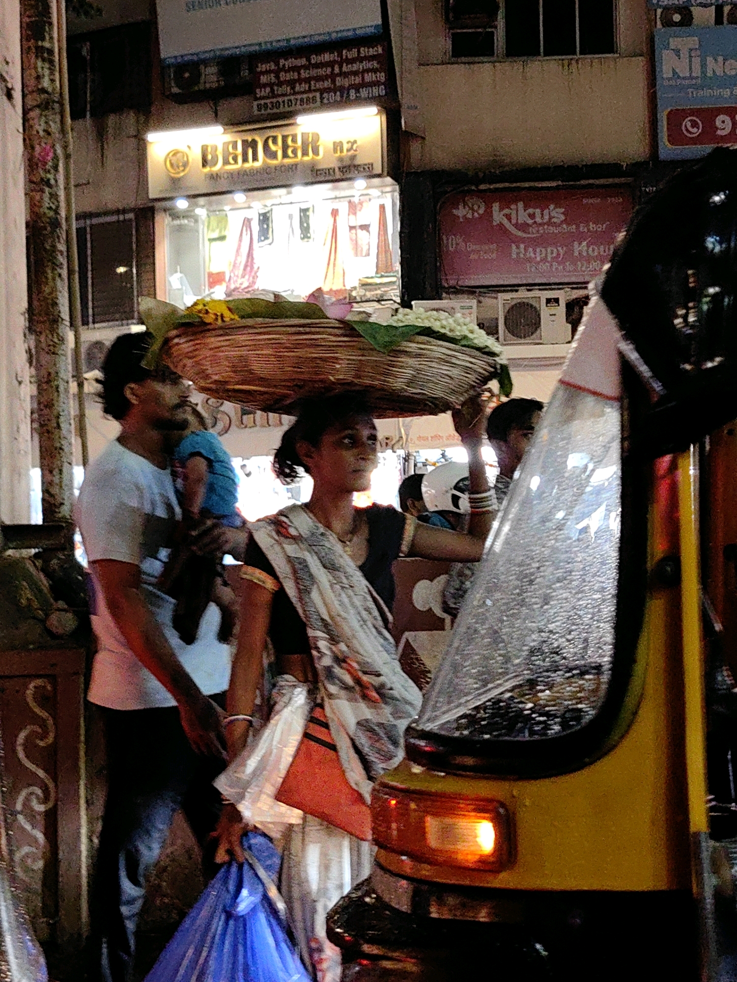 This is a nighttime street scene in Mumbai, India, likely in an area with shops and commercial activity. The foreground is dominated by the side of a yellow auto-rickshaw, with its orange turn signal illuminated. To the left of the auto-rickshaw, a woman is carrying a large basket of flowers and produce on her head. She is wearing a patterned sari. To her left, a man is holding a young child. Behind them, storefronts with illuminated signs are visible, including one for "BENCER" and another for "kilcu's restaurant & bar". The environment appears wet, with reflections on the ground, suggesting recent rain. People are dressed in typical Indian attire. The lighting is a mix of streetlights and shop signage, creating a somewhat hazy atmosphere.